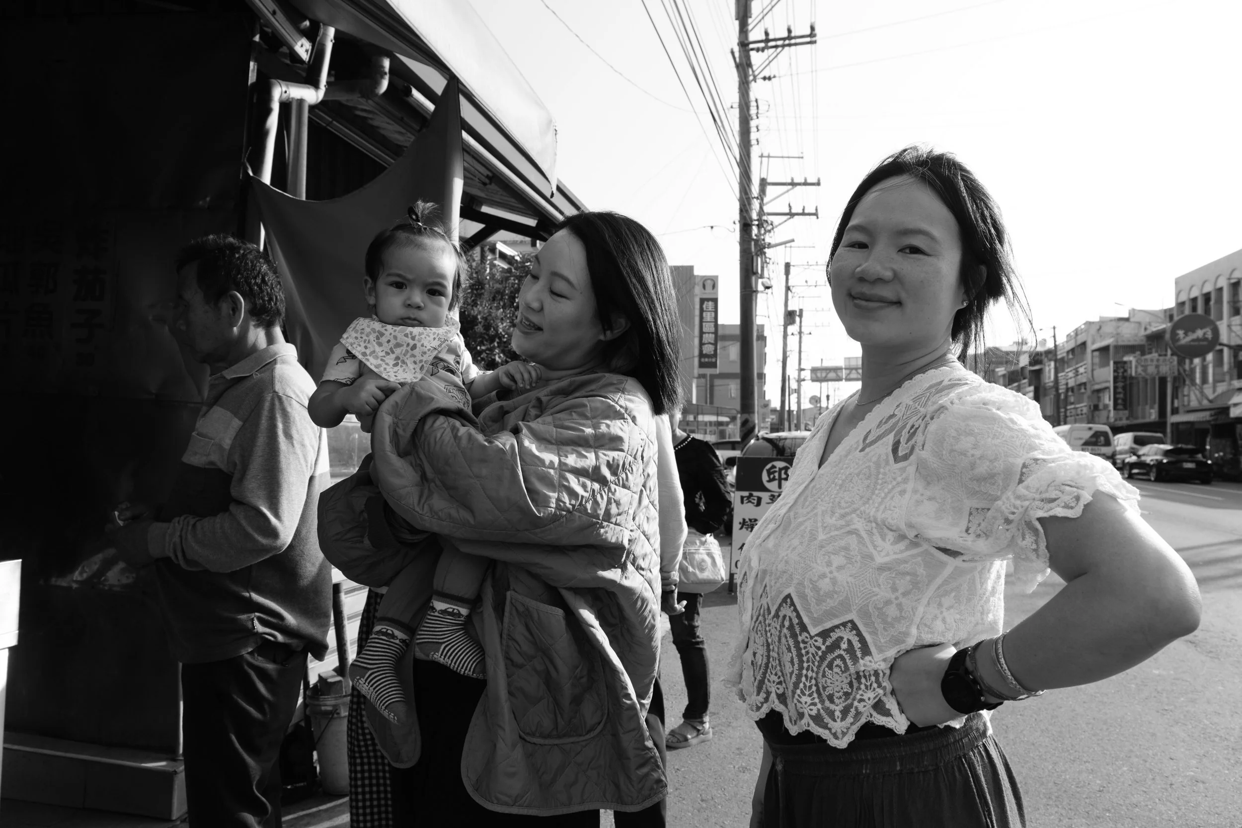 Two women are standing on a street in Tainan. One woman is holding a baby and looking at his face and the other woman is looking at the camera with a smile.