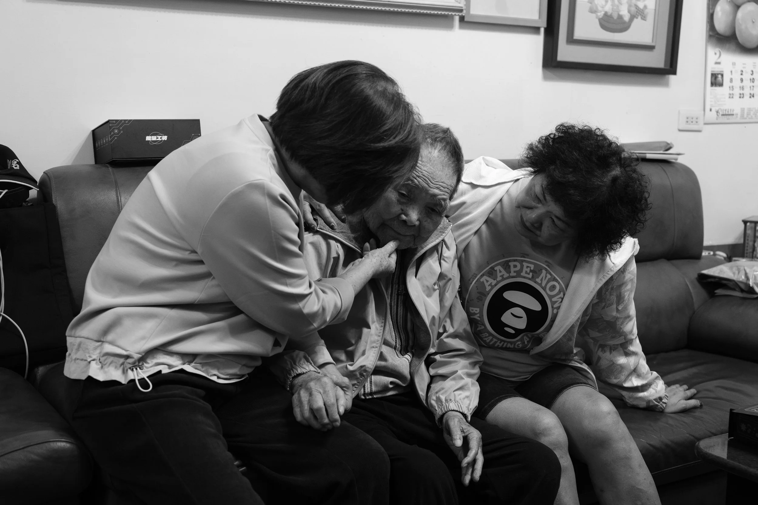 Two women are on either side of an old man sitting on a sofa in their ancestral home in Tainan, Taiwan. One of the women is touching the man's chin.