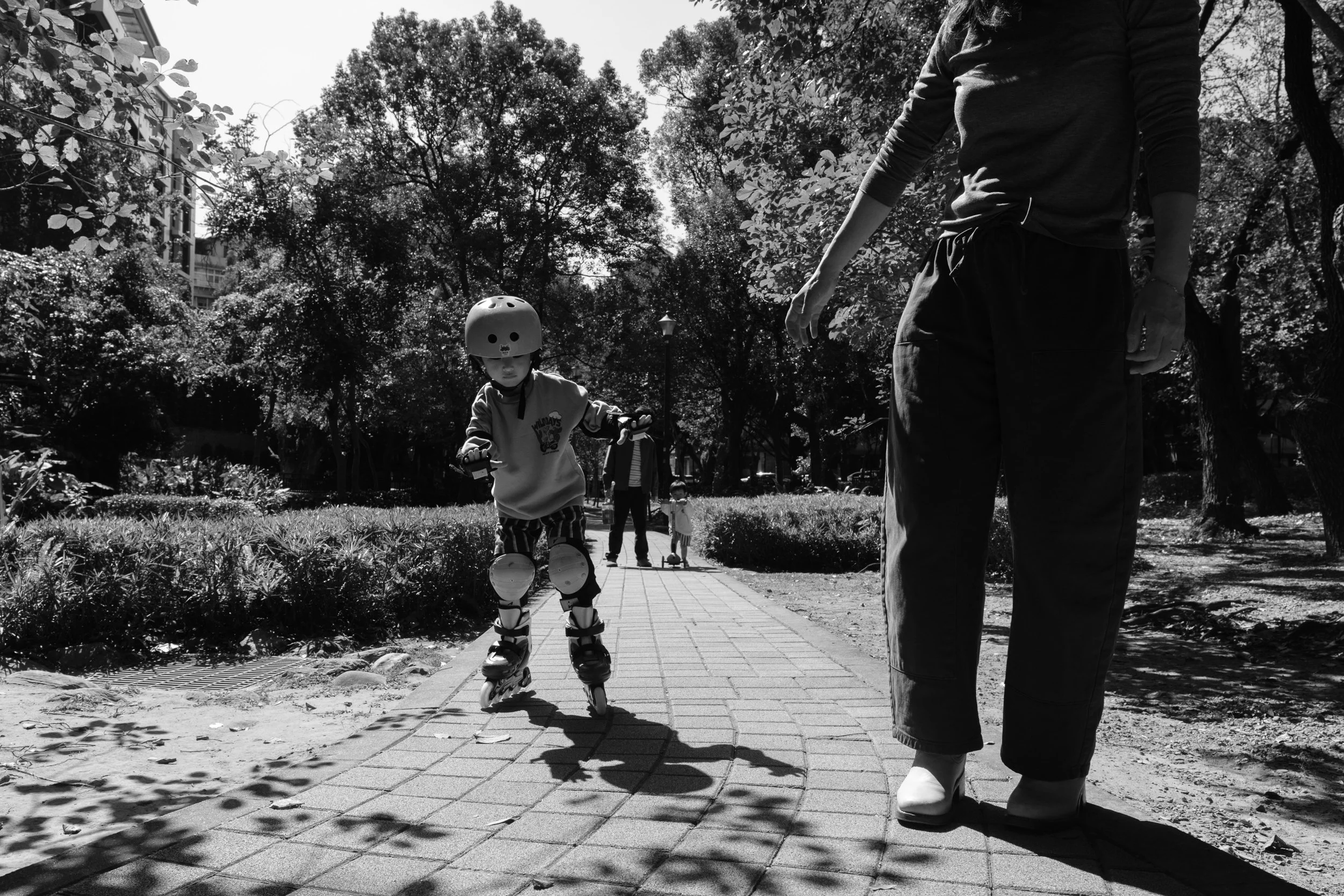 A child is rollerblading down a paved path in a park in Taipei during an outdoor documentary family photography session. His mom is waiting for him with an outstretched hand.