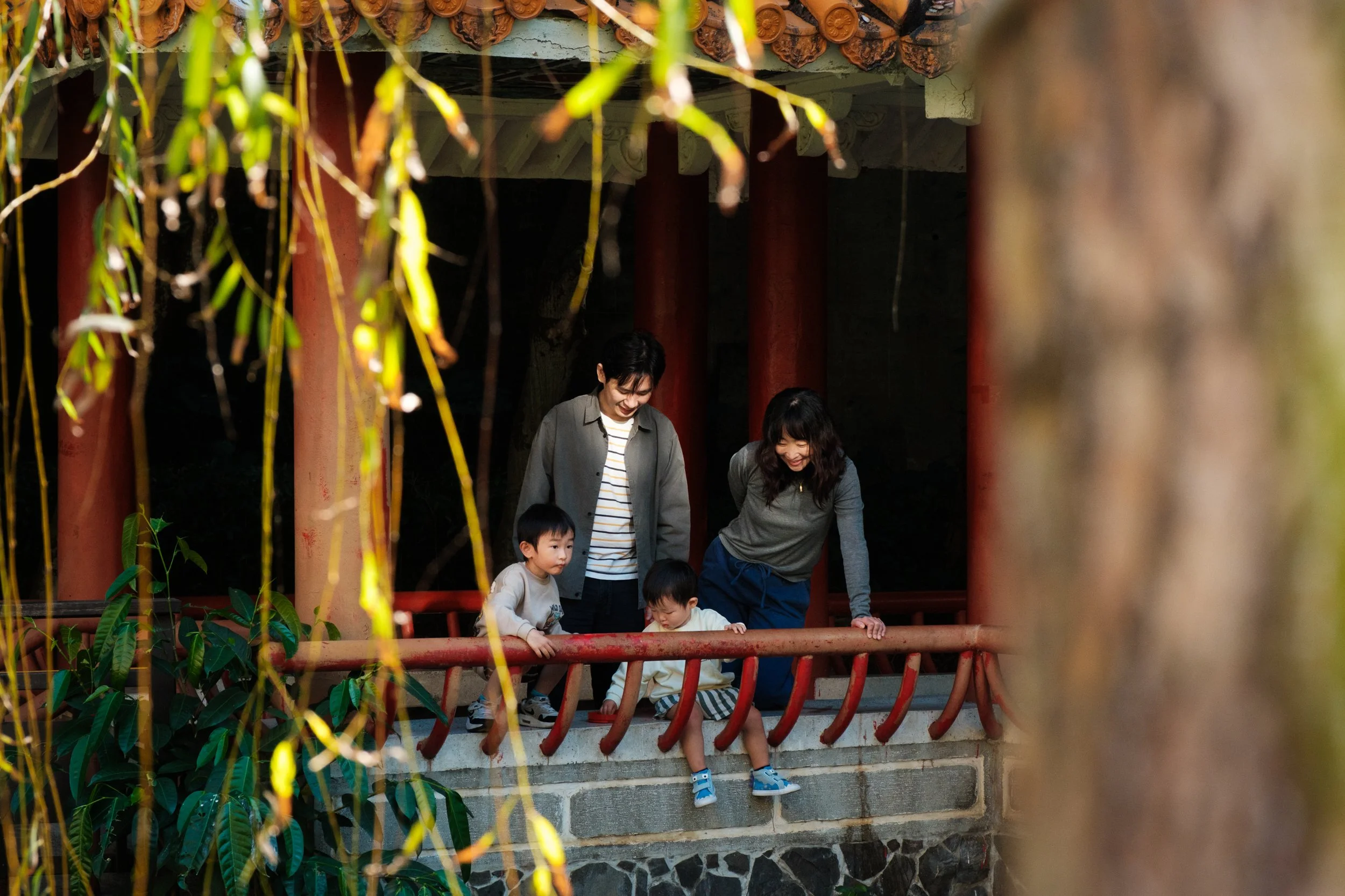 A family of four is standing under a pagoda in a Taipei park and looking at turtles in a pond below them during an outdoor Taipei family photography session. 