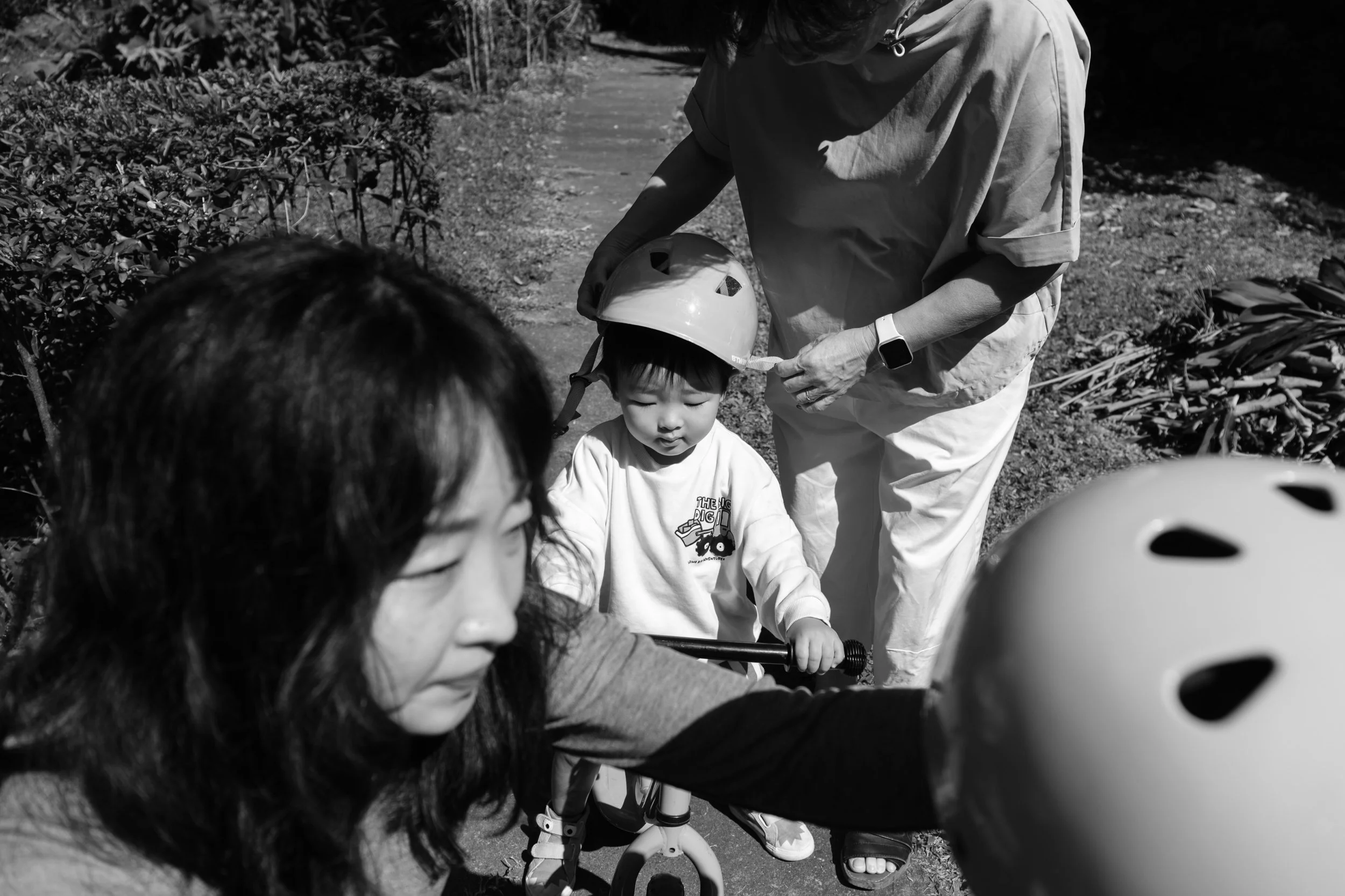 A mother and grandmother are putting helmets on two children in a park in Taipei, Taiwan. Photographed by Taipei photographer Cahleen Hudson during an outdoor family photoshoot.