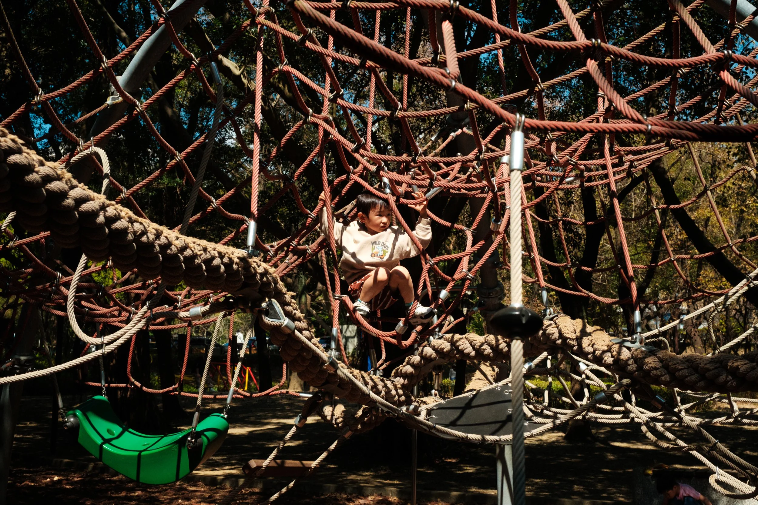 A little boy can be seen hanging in the middle of a rope jungle gym on a Taipei playground. Photographed by Cahleen Hudson during a documentary family photography session outside.