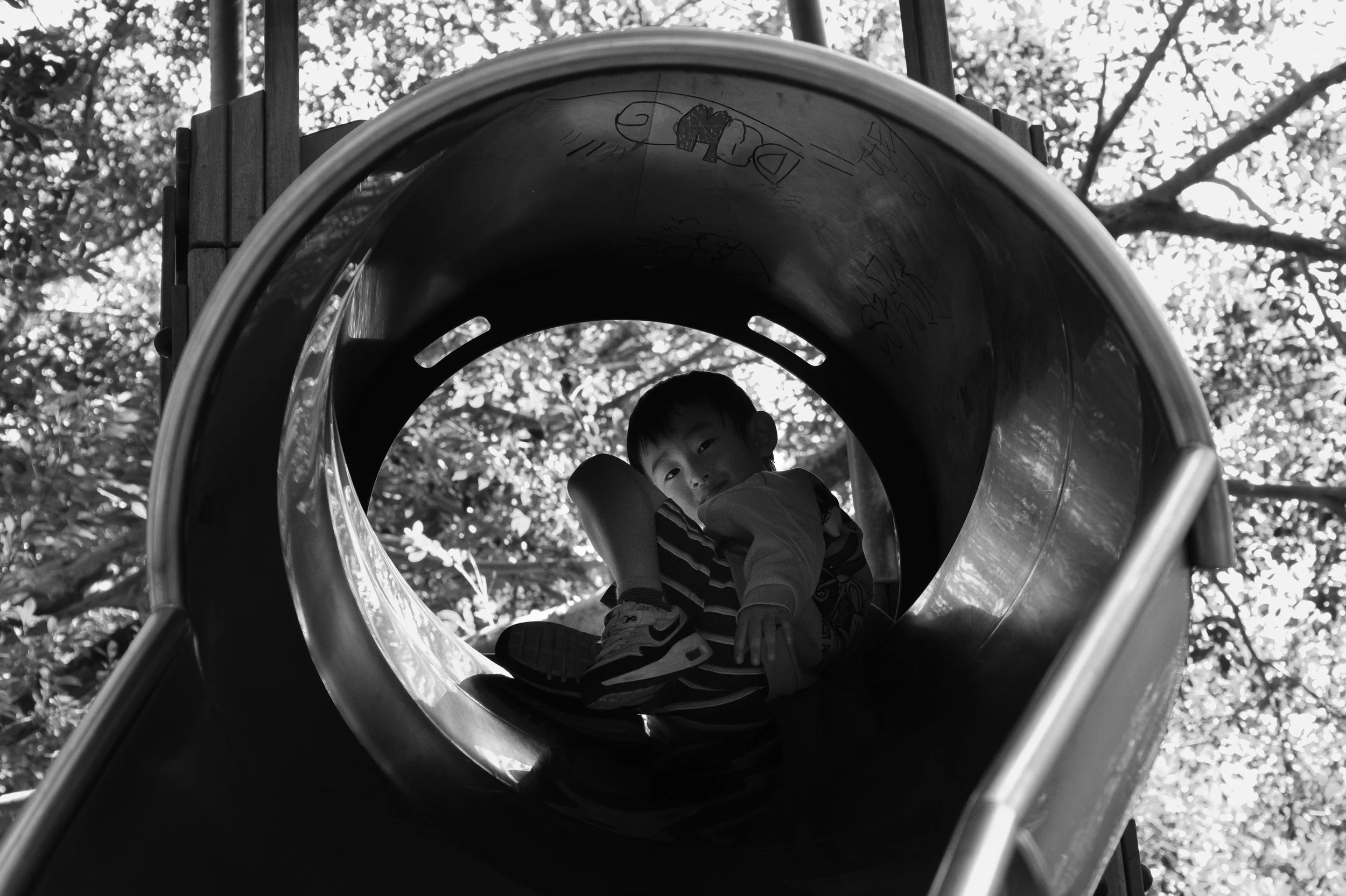 A boy is waiting at the top of a tube slide on a playground in Taiwan. He is being framed by the opening of the slide. Photographed during an outdoor family photoshoot in Tienmu.