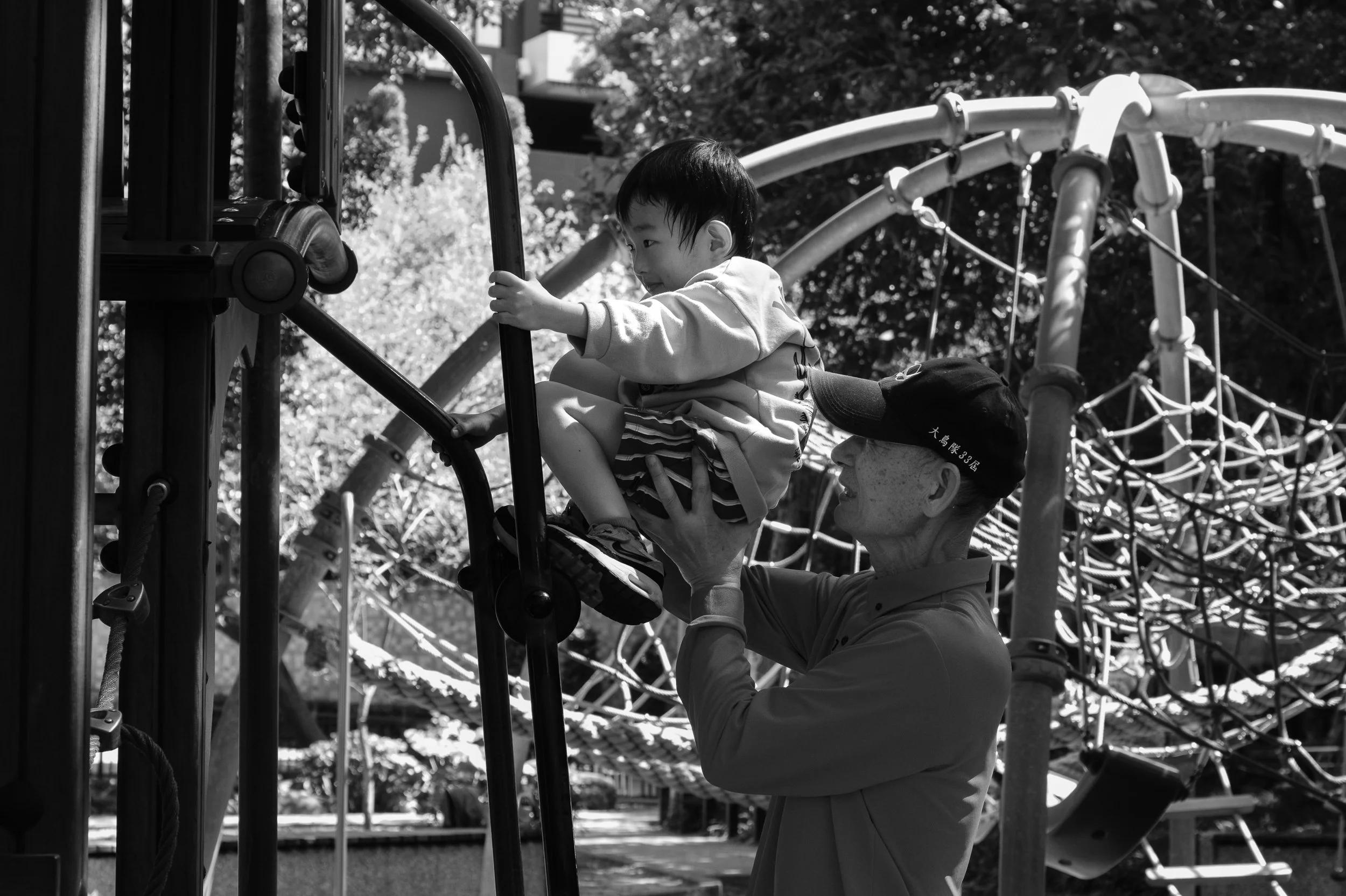 A boy's grandfather is helping him climb up playground equipment at a park in Tienmu, Taiwan during a Taipei family photoshoot outdoors.