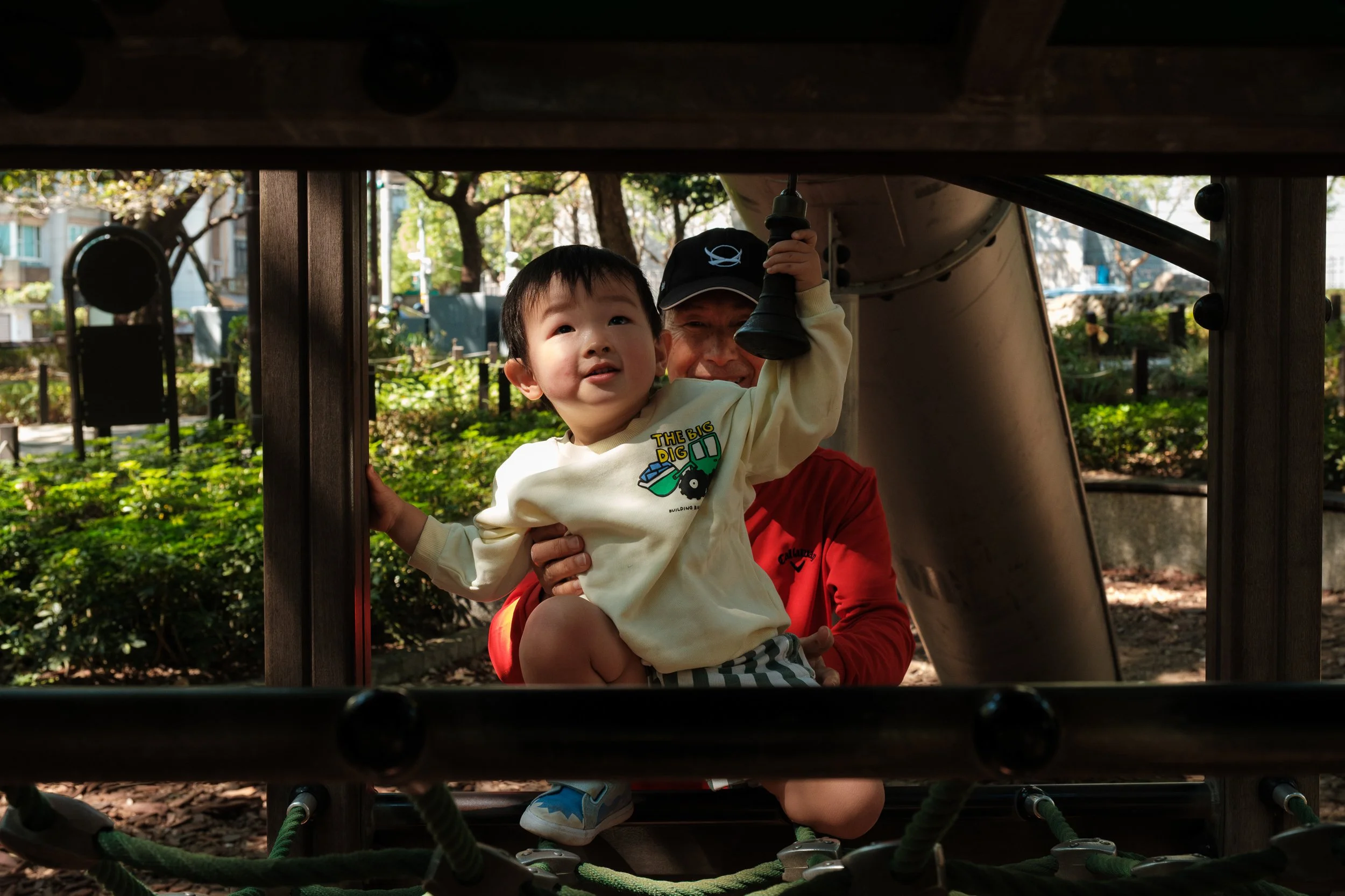 A little boy is climbing up playground equipment in a park in Taipei while his grandfather holds him from behind. Photographed by Taipei family photographer Cahleen Hudson.