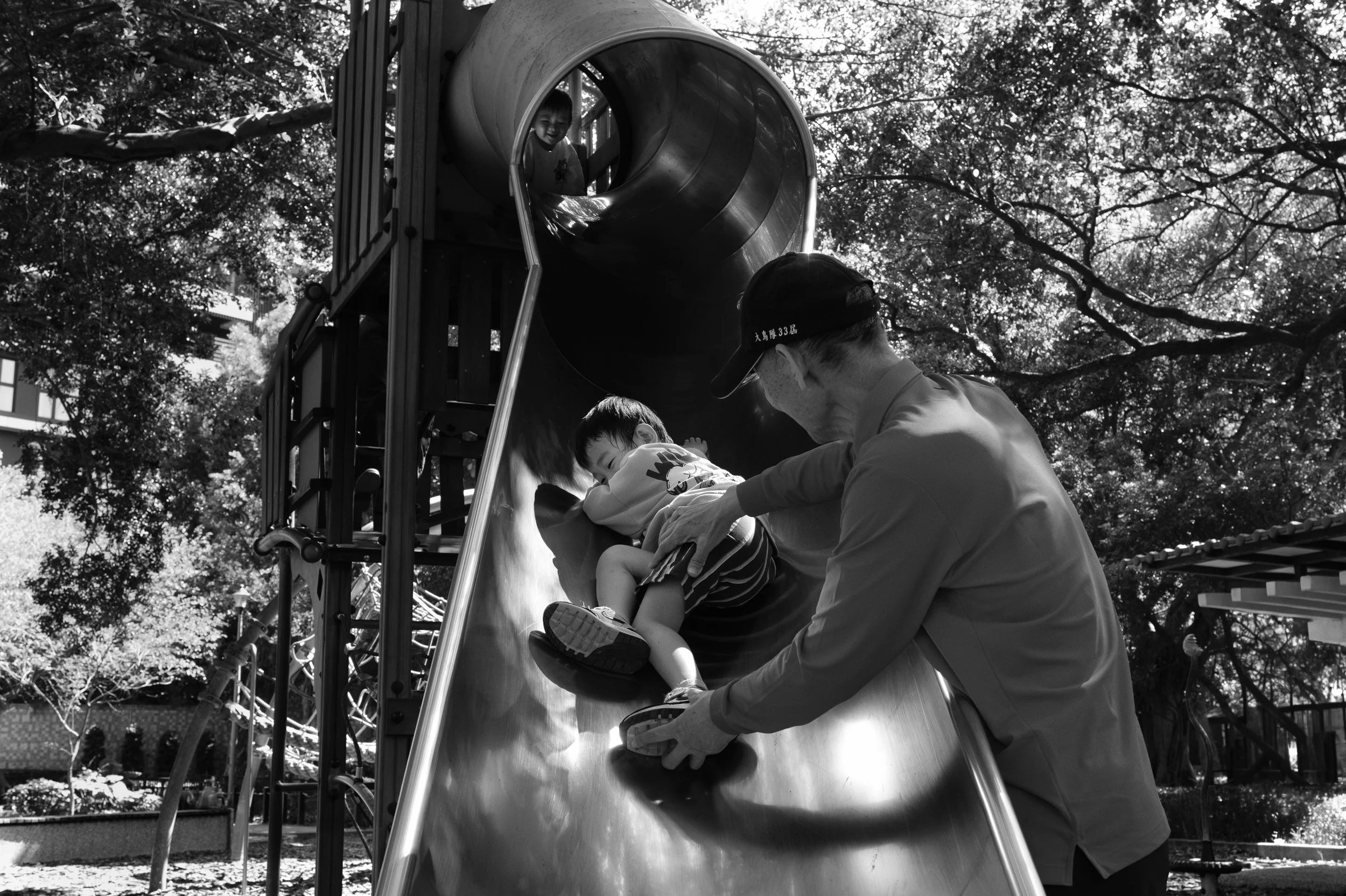 A child is sliding down a playground slide in Tienmu while his grandfather holds onto him. Photographed during an outdoor Taipei family photography session.