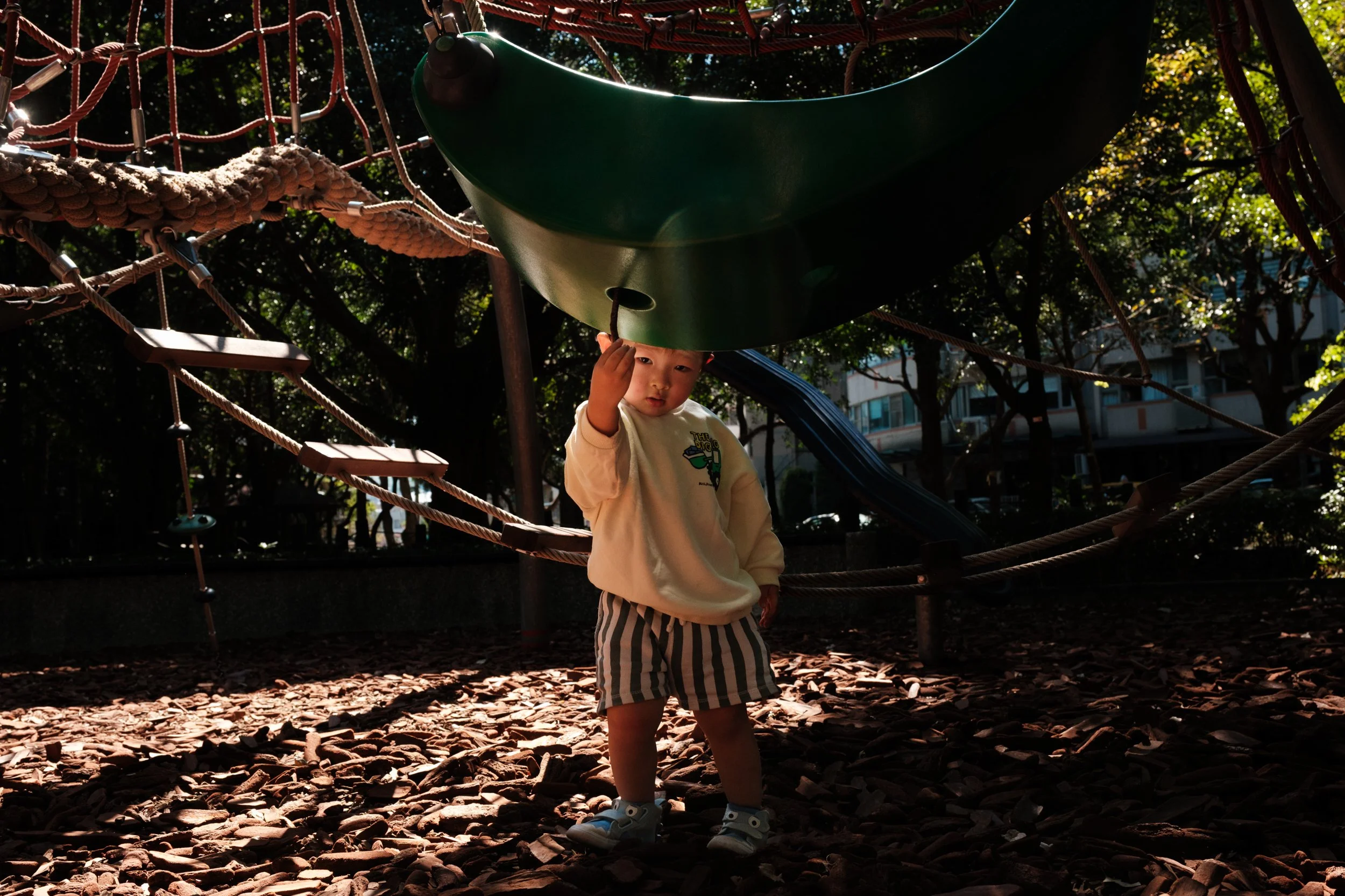 A little boy is peering under a swing on a playground in Taipei as natural light shines on his face. Photographed during an outdoor documentary family photography session in Taiwan.