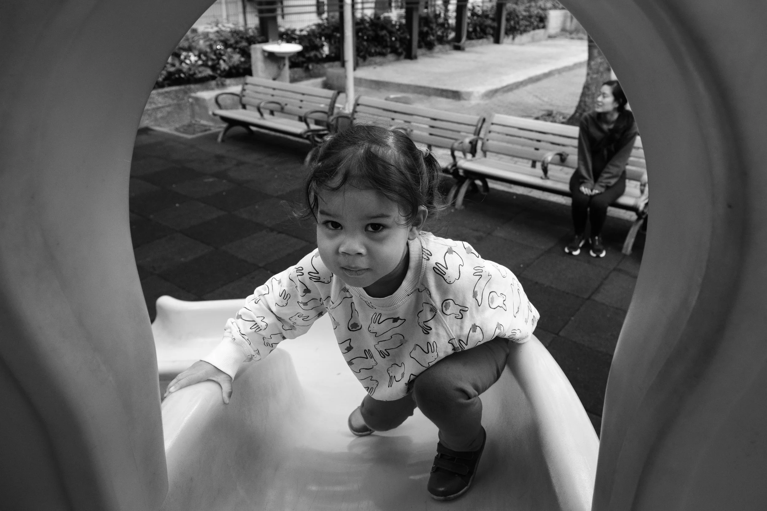 A little girl is climbing up a slide at a playground in Taipei during an outdoor family photoshoot. Her mom is sitting on a bench in the background.
