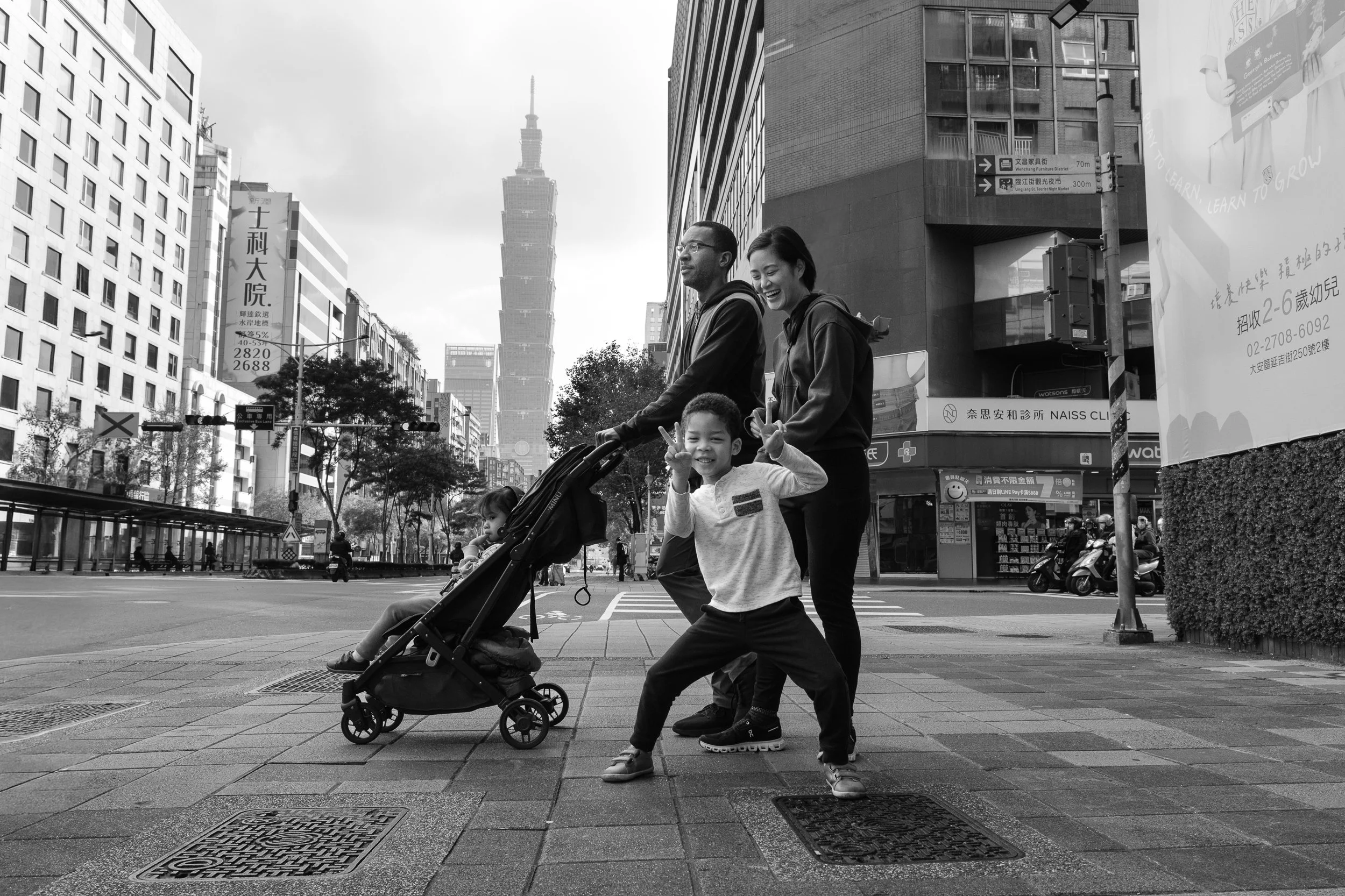 A family of four is waiting to cross a Taipei street during an outdoor documentary family photography session. Taipei 101 can be seen in the background.