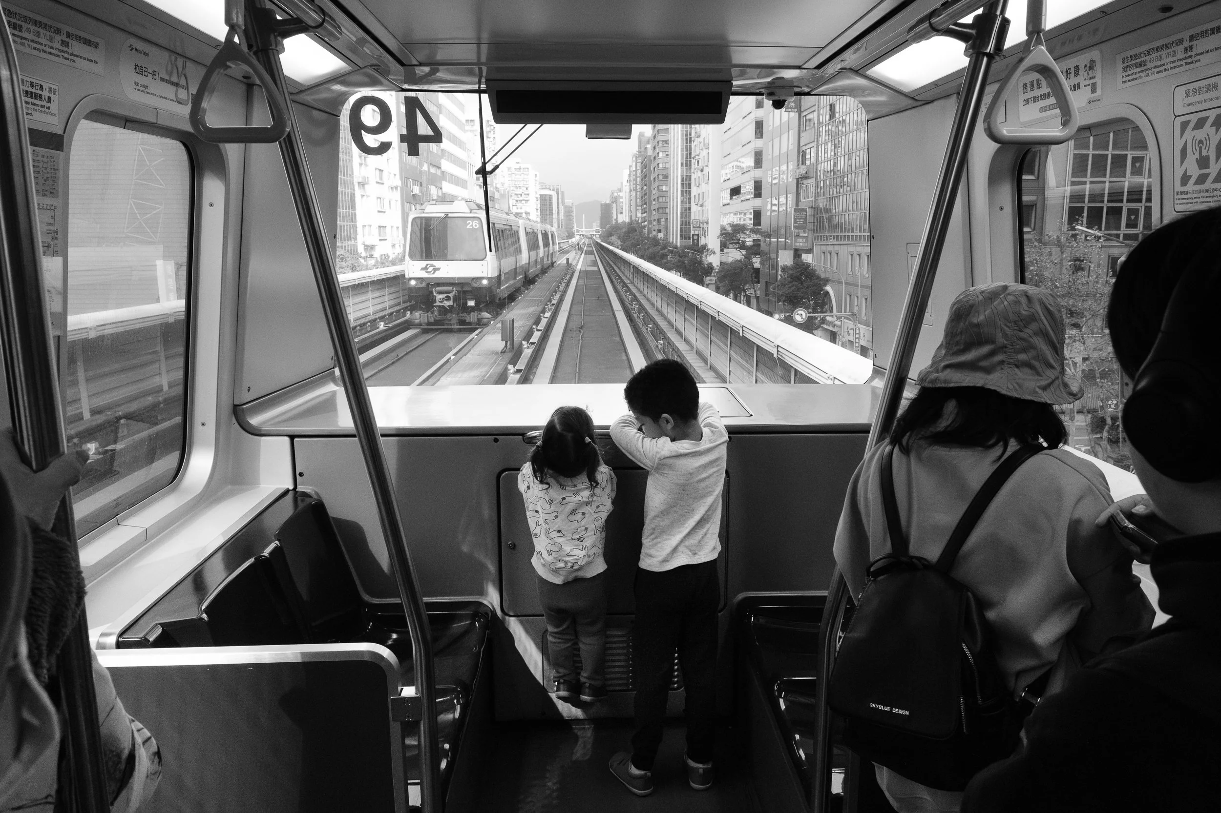 A brother and sister are riding the brown line of the Taipei MRT and the city can be seen below them through the window.