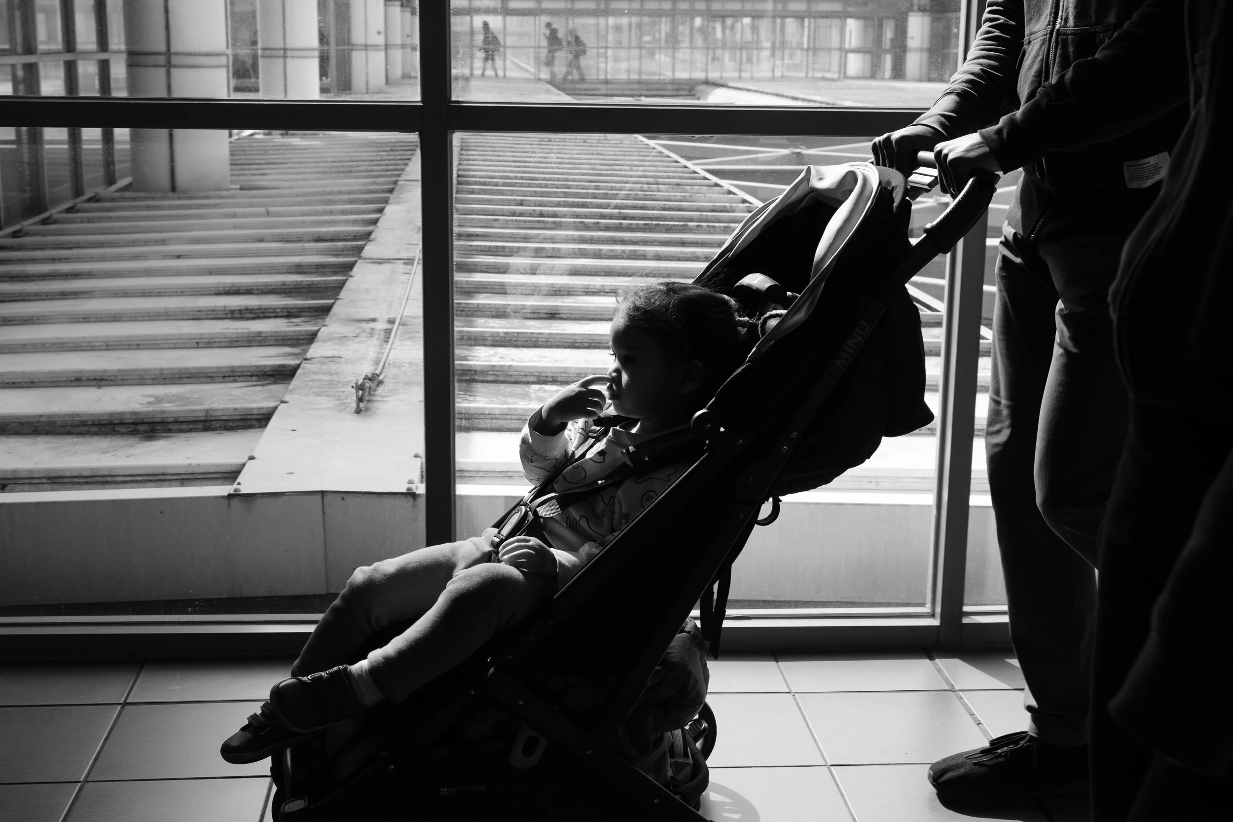 A little girl is being pushed in her stroller at a Taipei MRT station and his touching her nose with her finger. A big window is behind her.