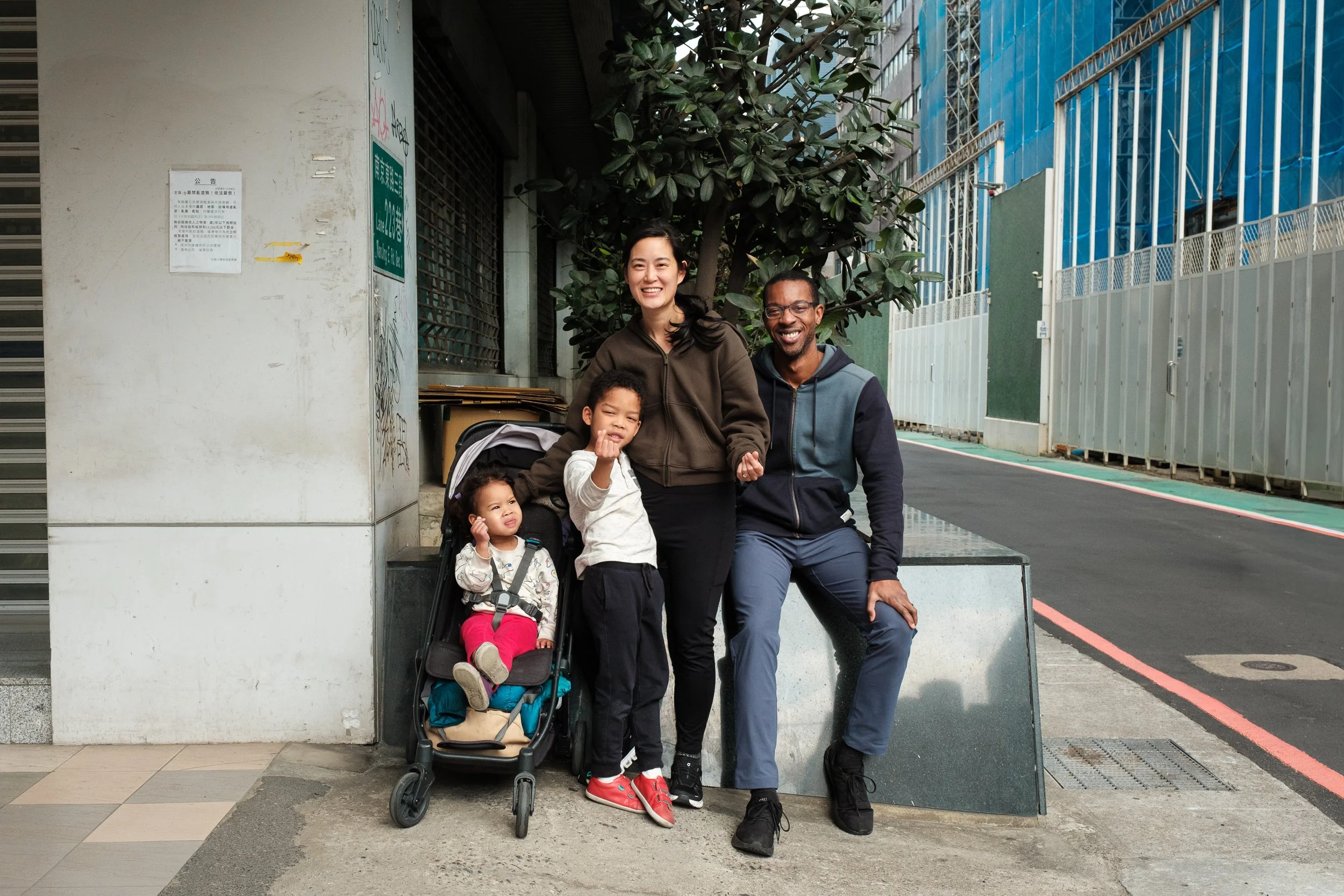 A family of four is sitting on a planter and using their fingertips to make a heart sign during a Taipei family photoshoot. 
