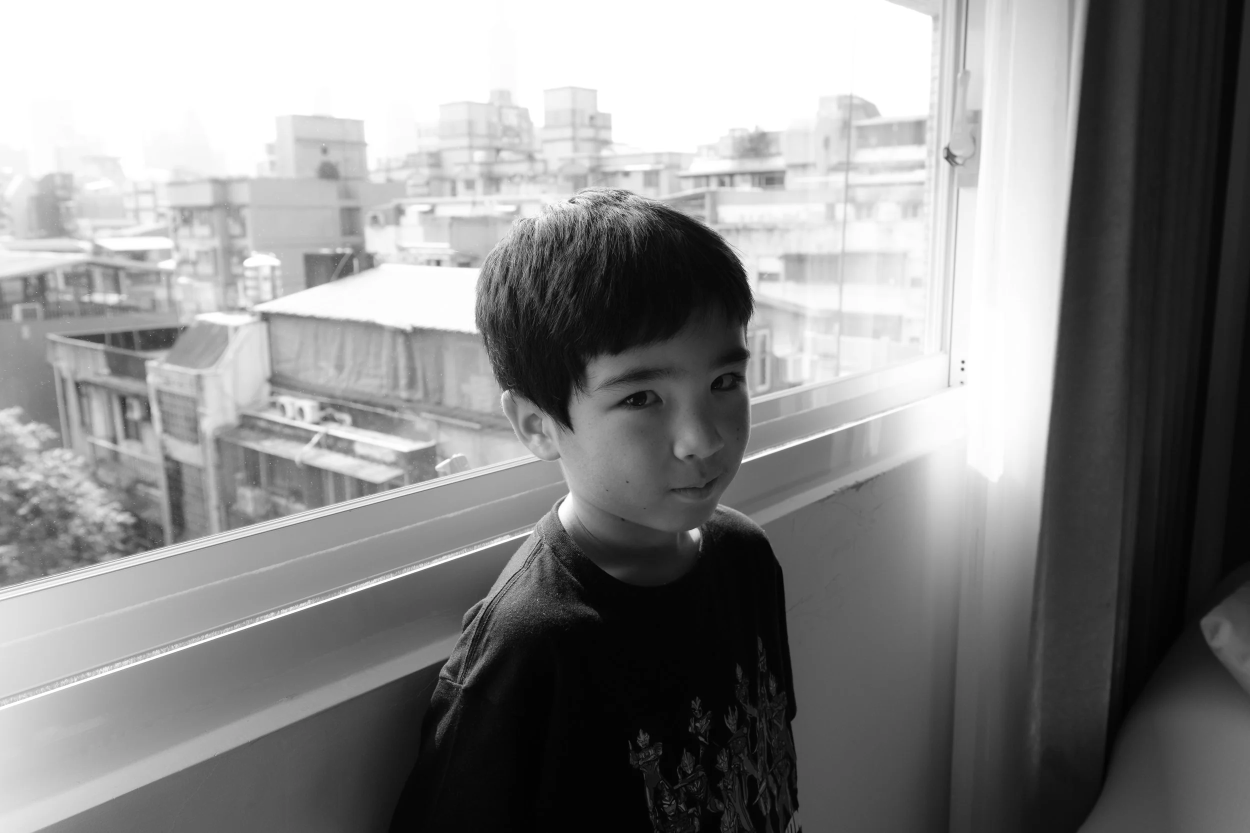 A little boy is standing in front of a window in a Taipei apartment; a city scene can be seen through the window.