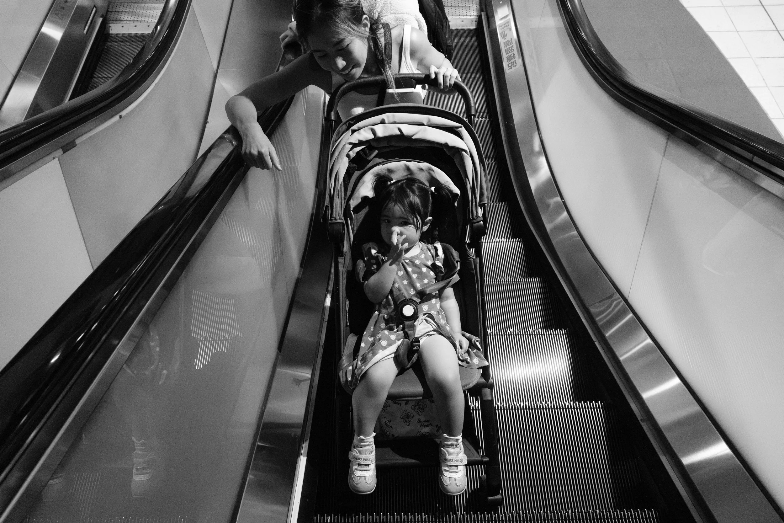 A mother takes her little girl in a stroller up an escalator in a Taipei MRT station. She's leaning over and looking at her child. 