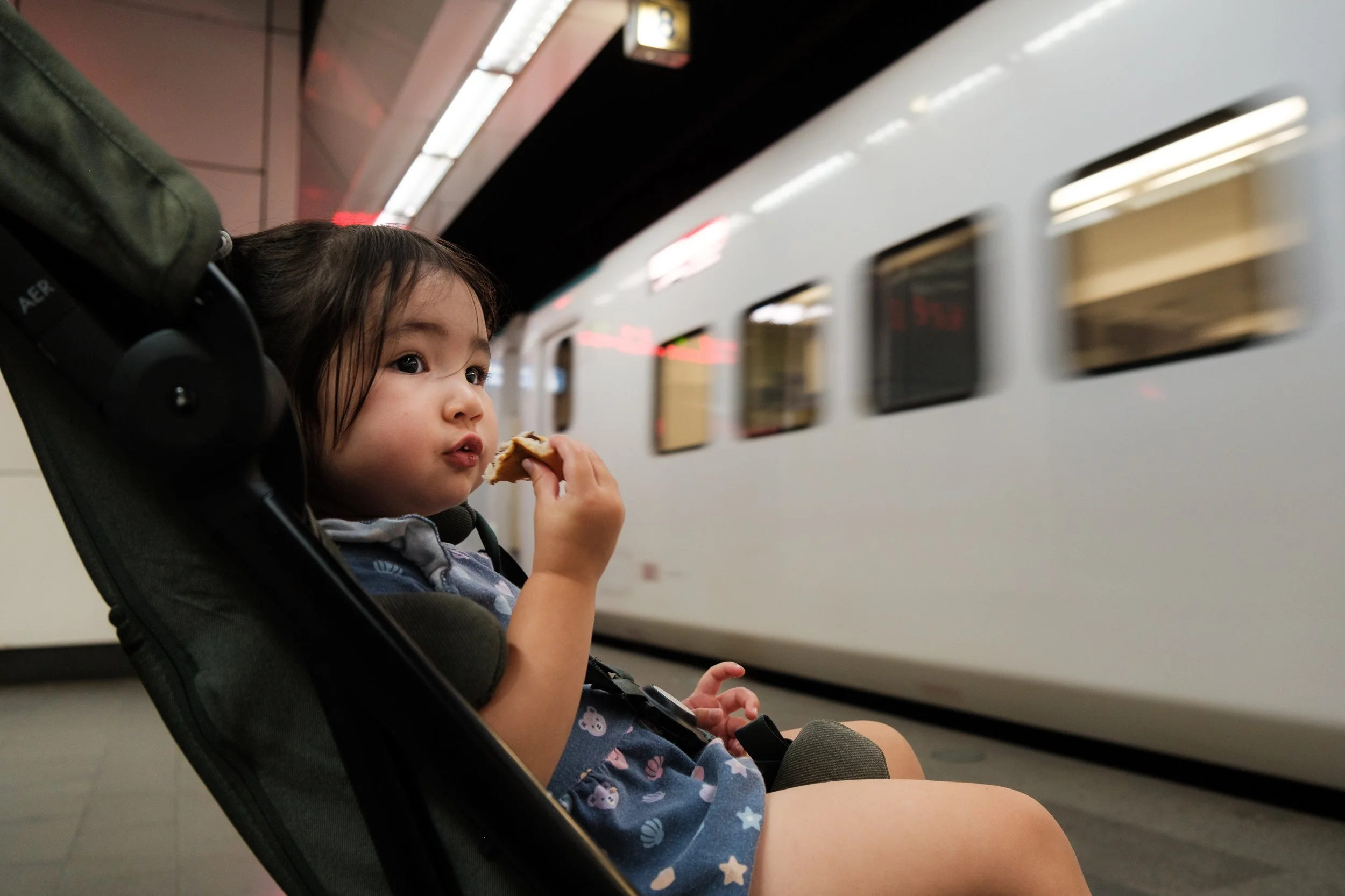 A little girl is sitting in her stroller and eating a snack as train pulls up to the platform at a Taipei train station. 