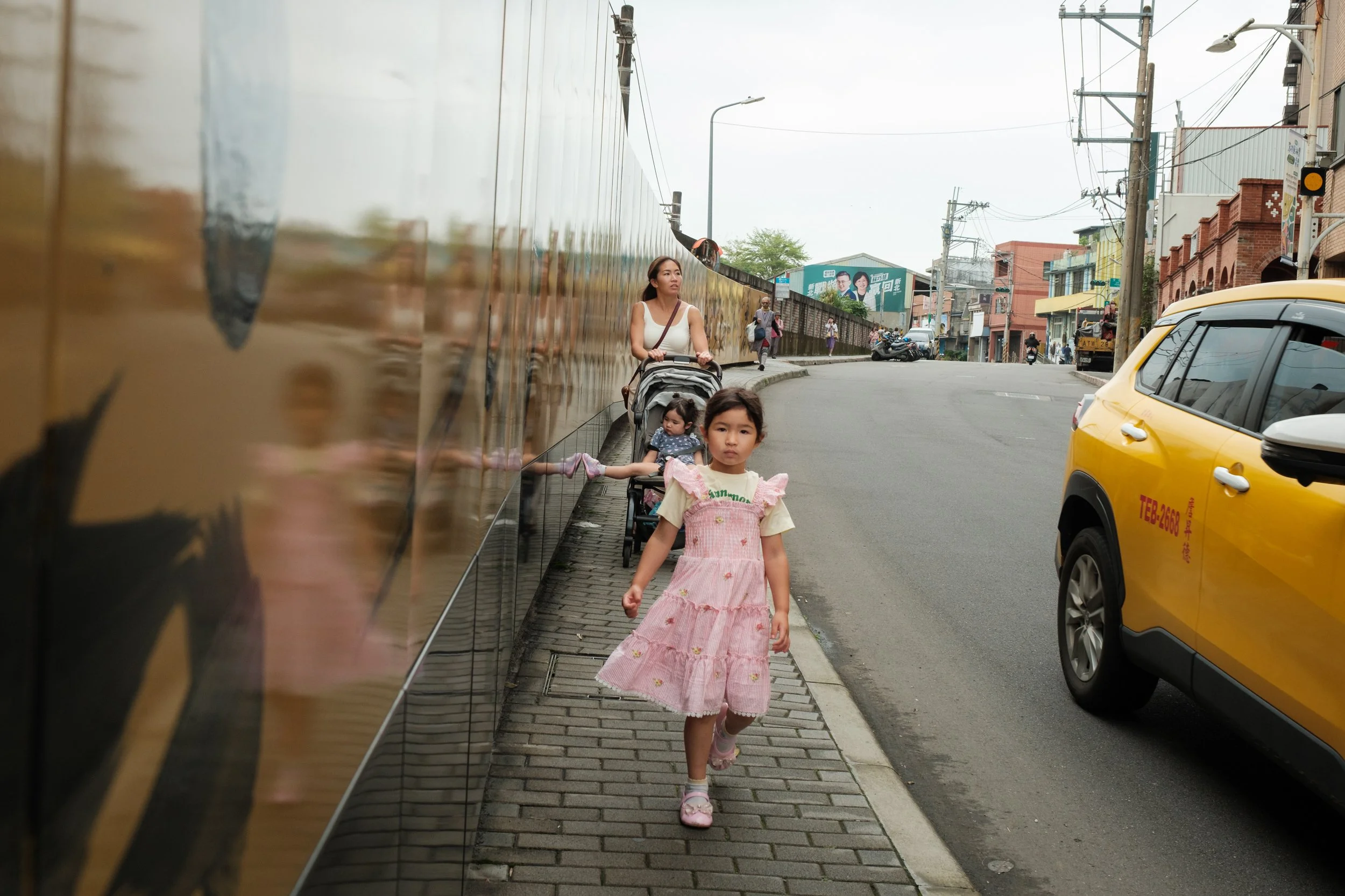 A young girl walks down a street in Yingge. A taxi is passing her and her mother is in the background. Photographed during an outdoor family photoshoot in Taiwan. 