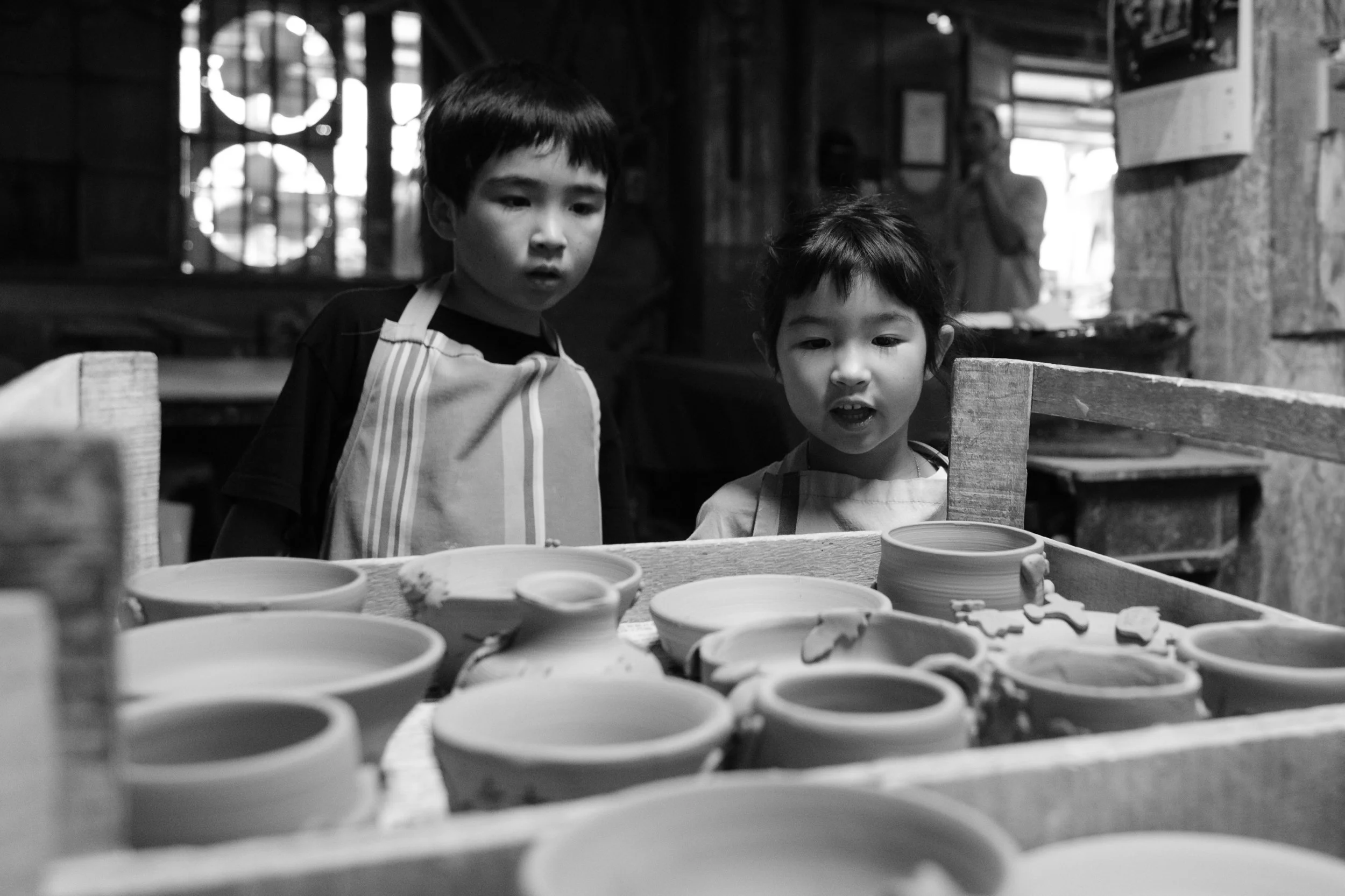 Two children are looking at rows of clay pots drying in an Yingge DIY ceramics shop. Photographed during an outdoor family photography session by taipei photographer Cahleen Hudson.