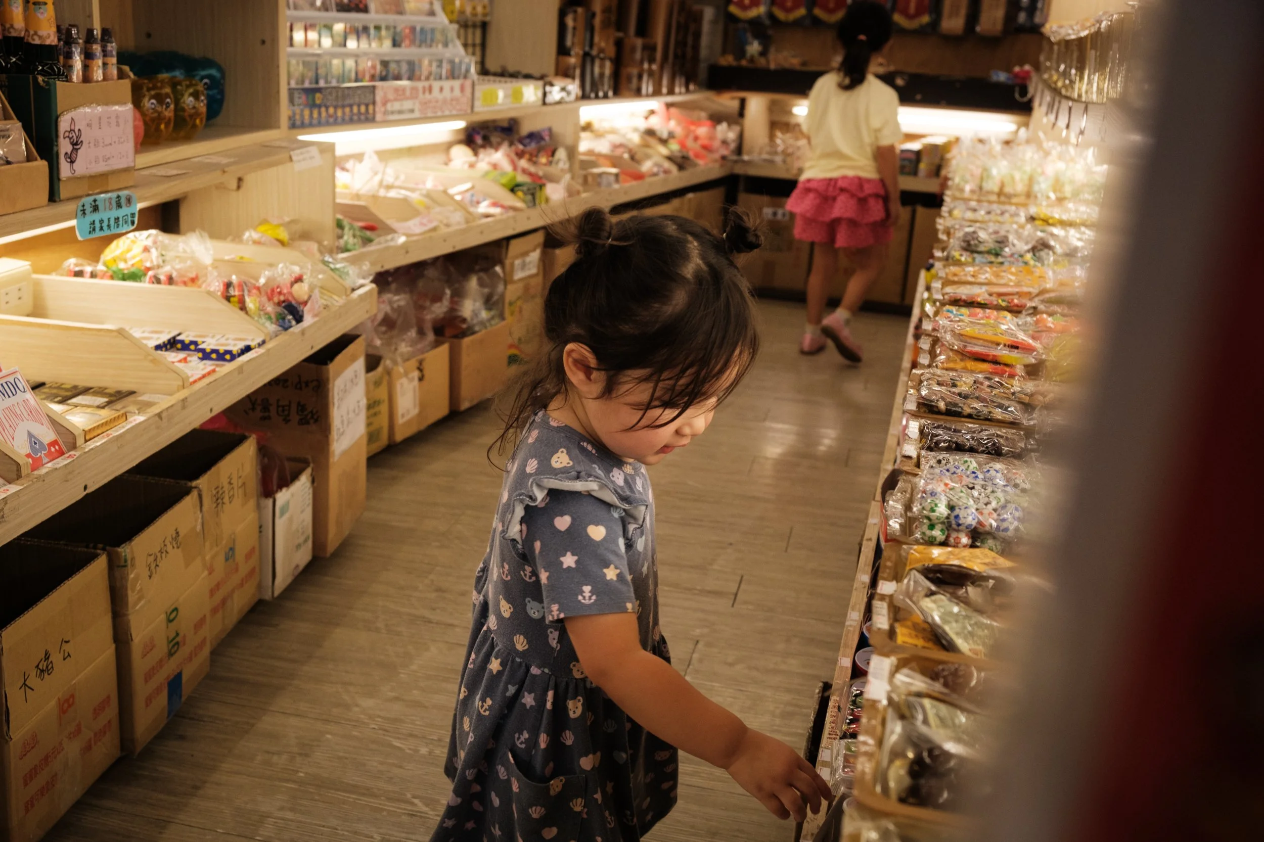 A little girl is looking at candy in a shop on Yingge Old Street in taiwan. Her big sister is in the background. Photographed during a family photoshoot by Cahleen Hudson.