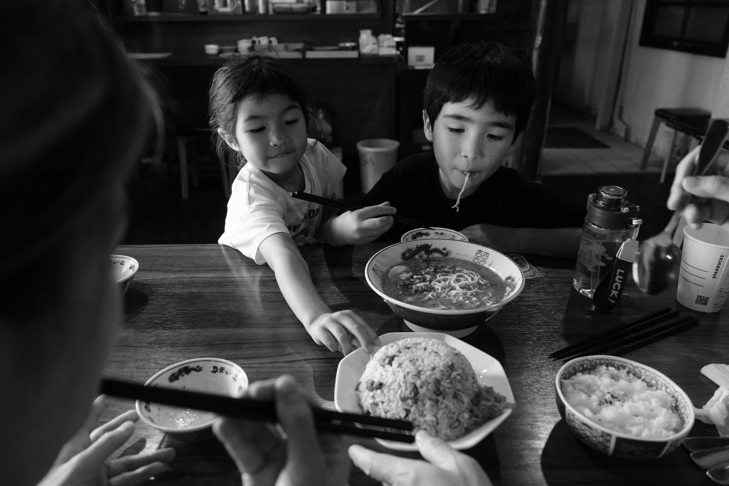 Two kids are reaching for their food in a resturant in Yingge, Taiwan. The little boy is slurping up a noodle that's hanging out of his mouth. 
