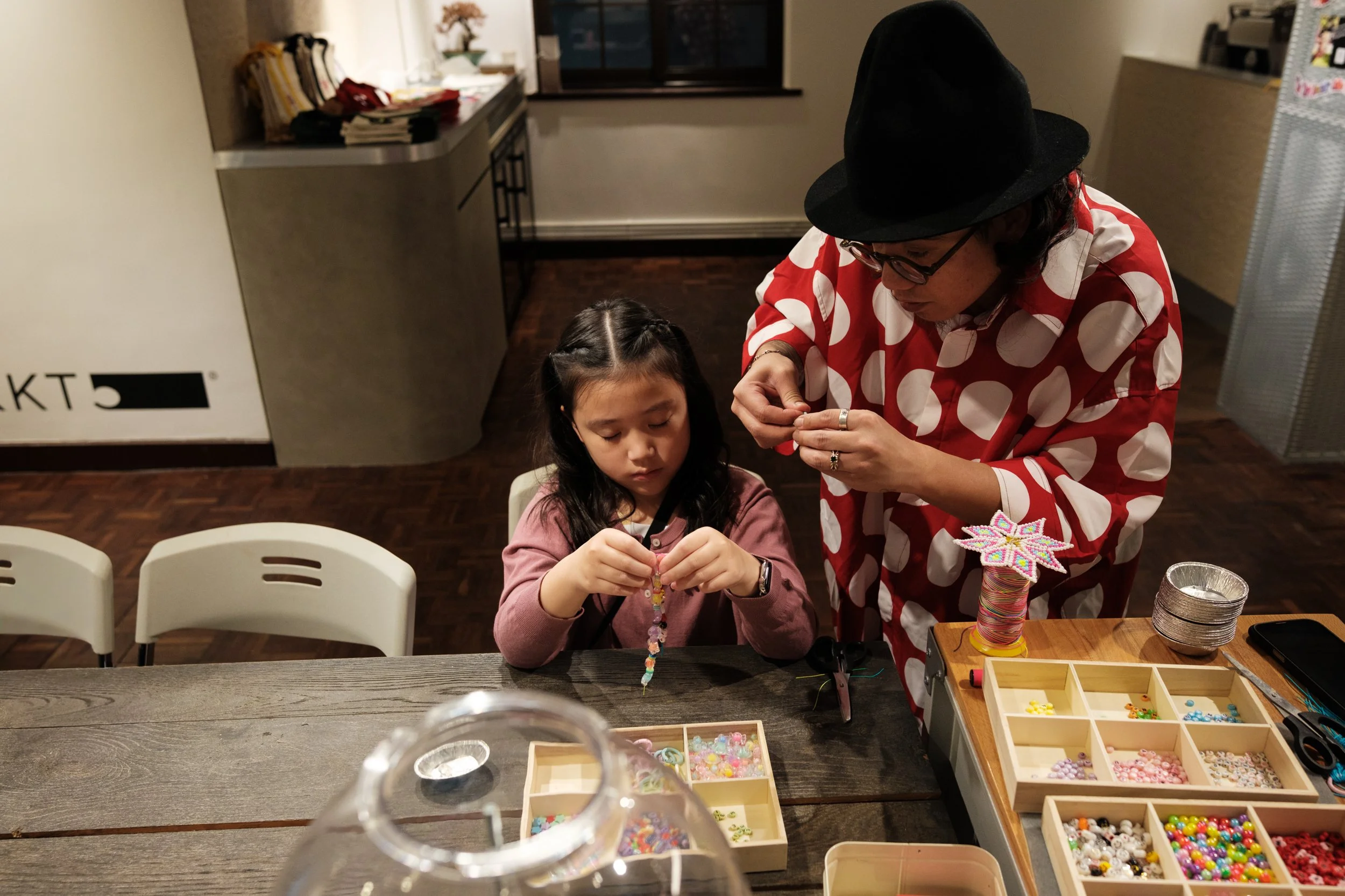 A man is next to a little girl at big wooden table and helping her make a bracelet at a beading station at a child's birthday party in Taipei, Taiwan.