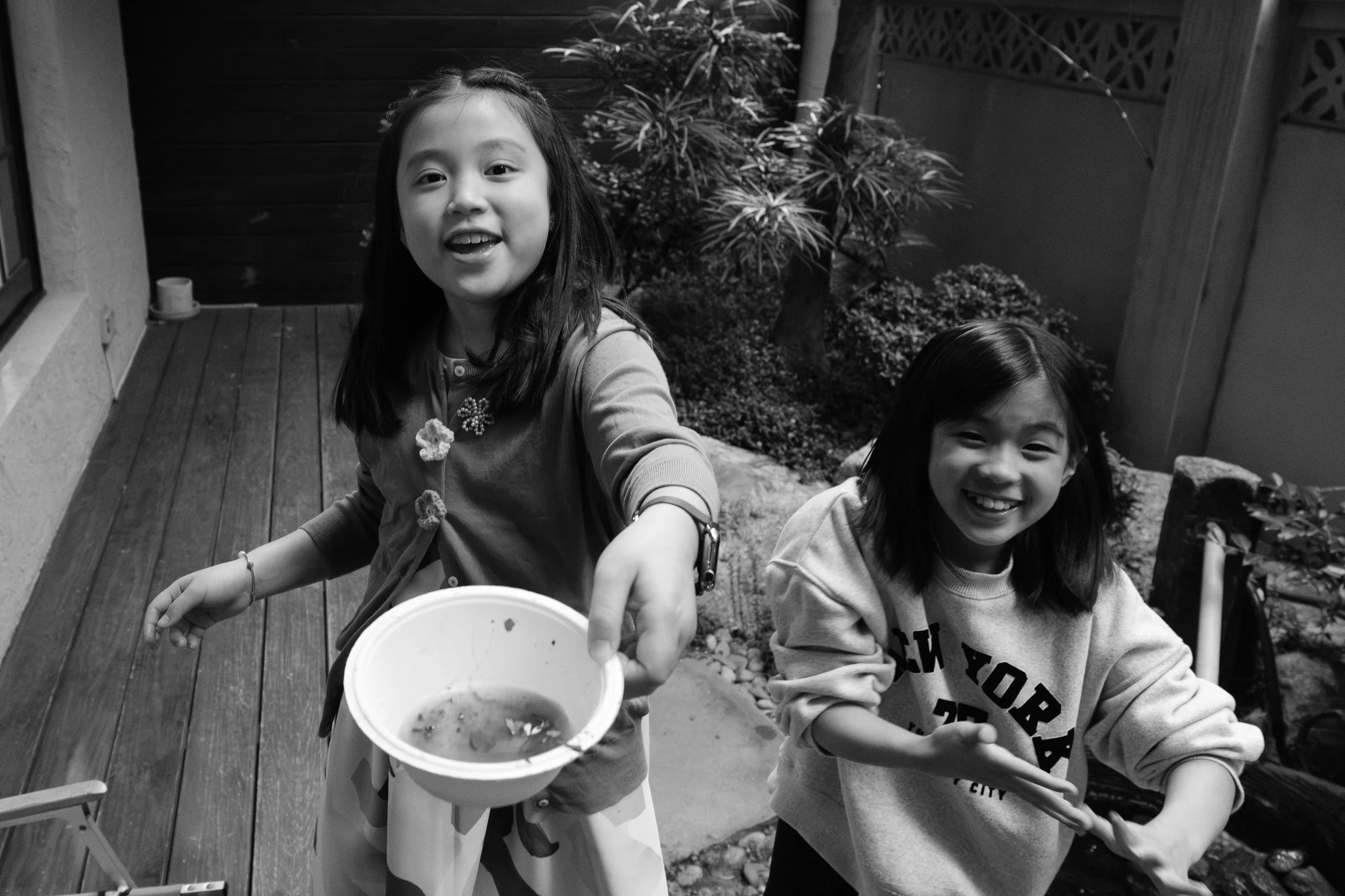 Two young girls are laughing as they serve someone a paper bowl of crushed up leaves. They are pretending to cool on the patio of MRKT5 in Taipei during a child's birthday party.