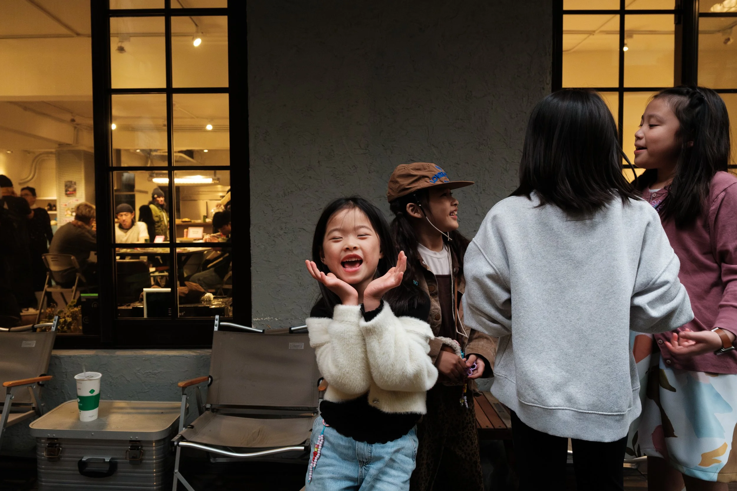 A little girl has her hands on her face and is laughing with her friends on the patio of MRKT5 during her friend's birthday party in Taipei. People can be seen inside through the windows behind her.
