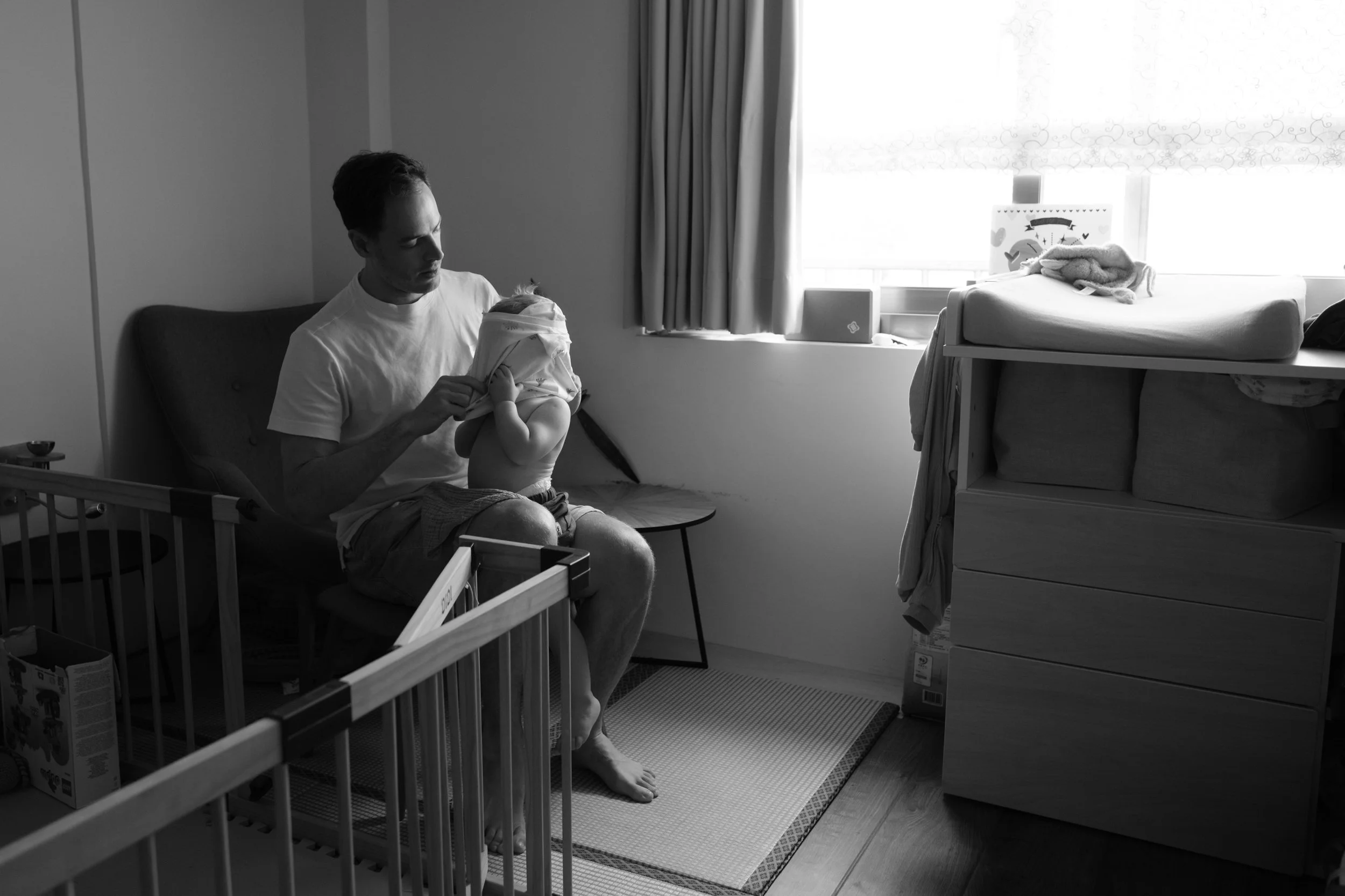A father puts a shirt on his toddler son in front of a big window in their Taipei apartment during and indoor family photoshoot.