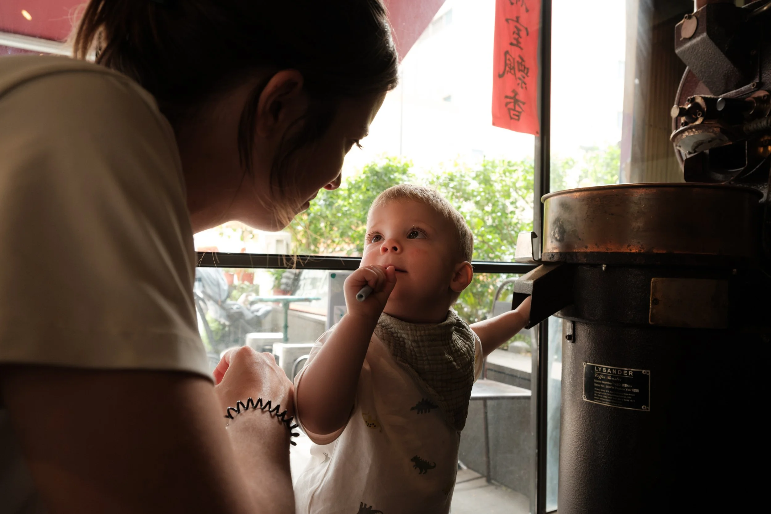 A little boy is looking up at his mother as he touches a coffee roaster in a Taipei coffee shop during a family photoshoot.