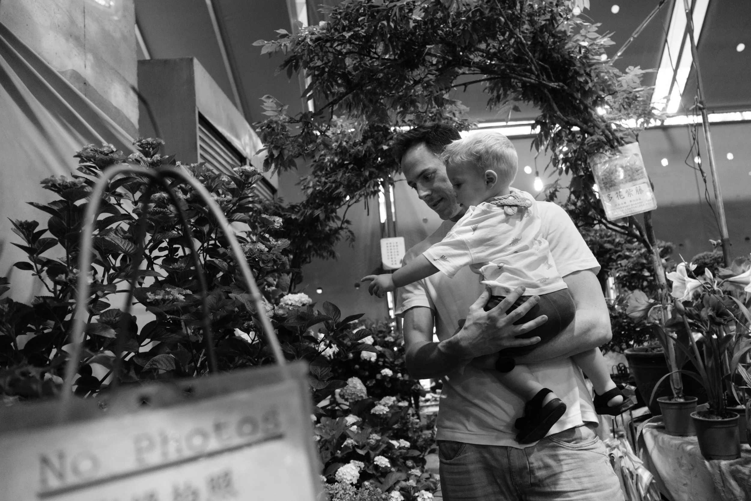 A man is holding his toddler son at the Jianguo Flower market in Taipei during an outdoor family photography session.
