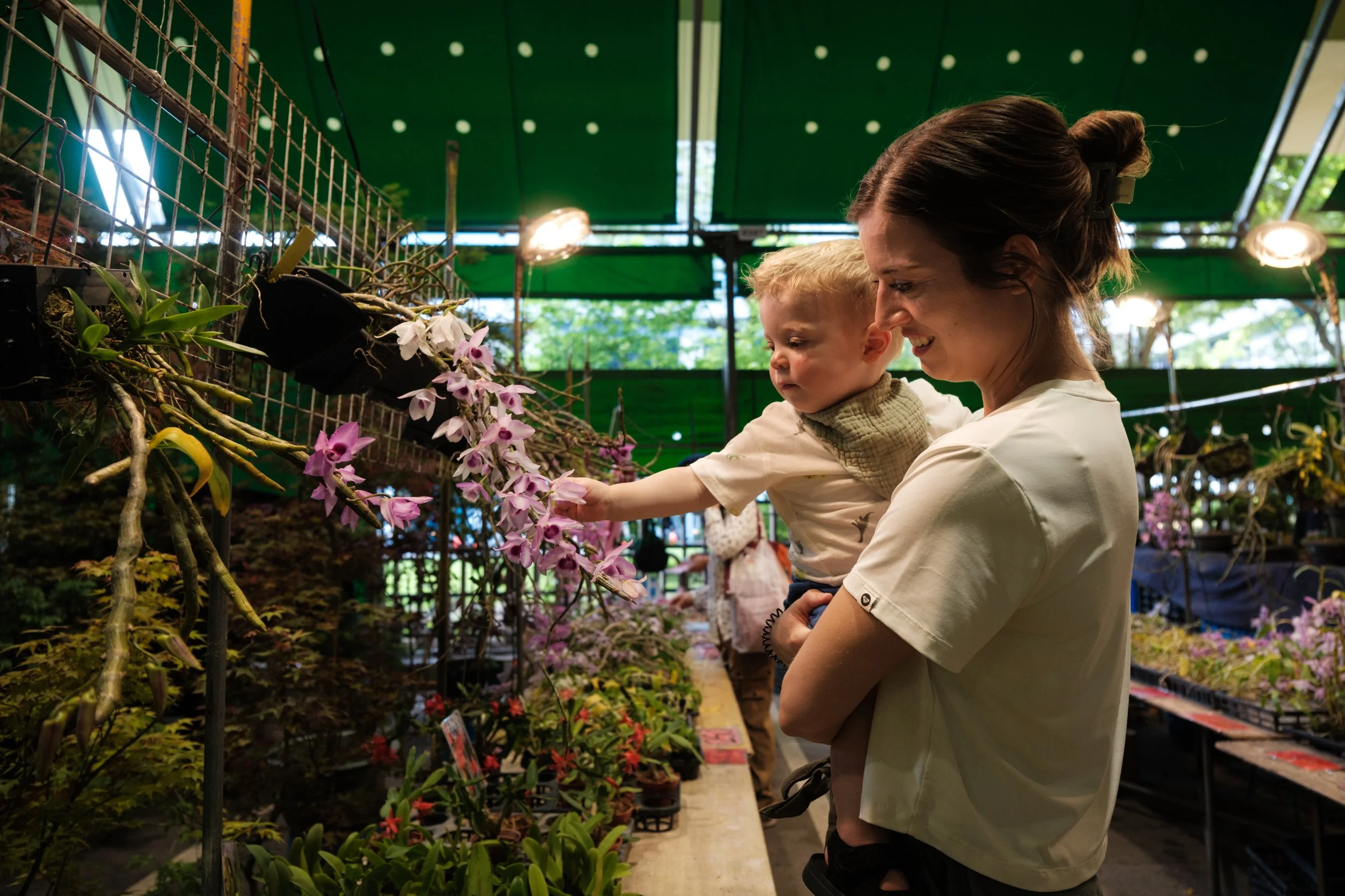 A woman holds her toddler son and helps him touch plants at the Jianguo Flower Market in Taipei during an outdoor documentary family photography session.
