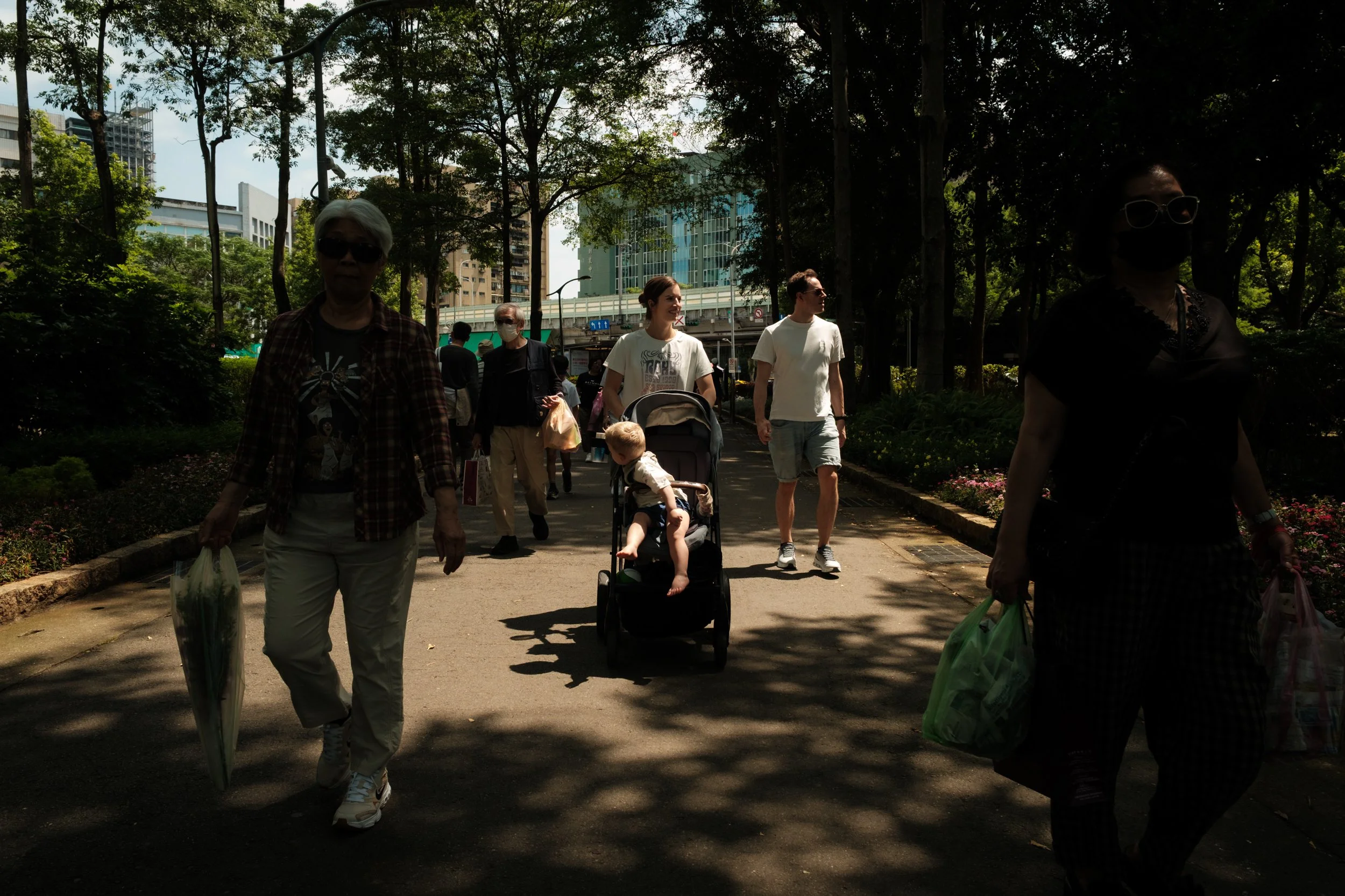 A couple is oushing their son in a stroller on a sunny path in Daan Park during an outdoor family photoshoot.
