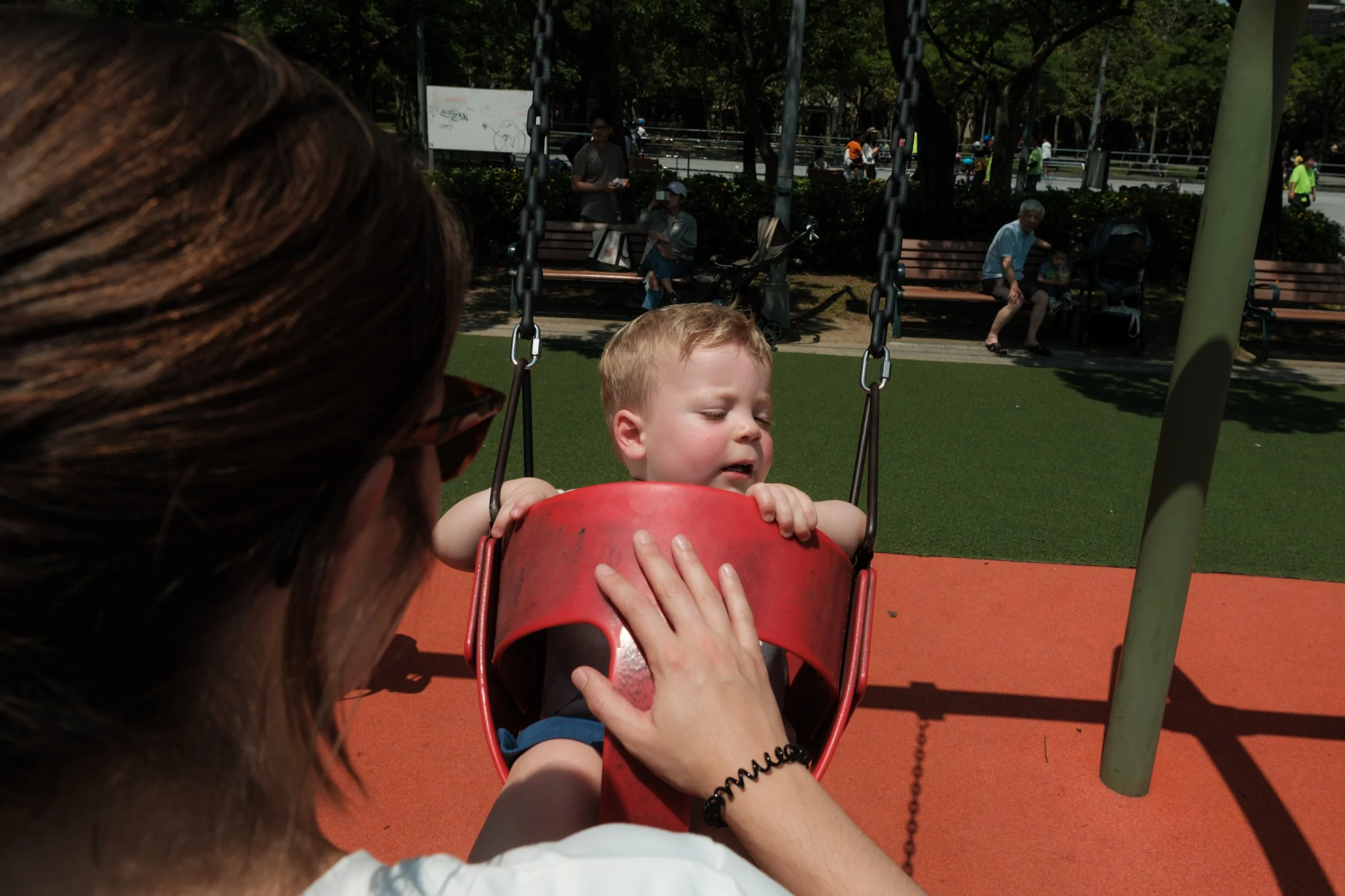 A mother is pushing her toddler son in a swing on the playground at Daan Park during an outdoor Taipei family photography session.