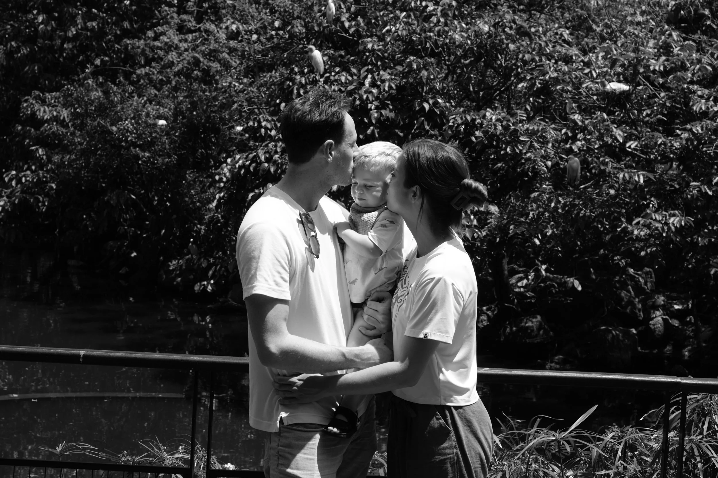 A mother and father are holding their toddler son between them and each kissing his cheek at Daan Park in Taipei, Taiwan during a documentary family photography session outside.