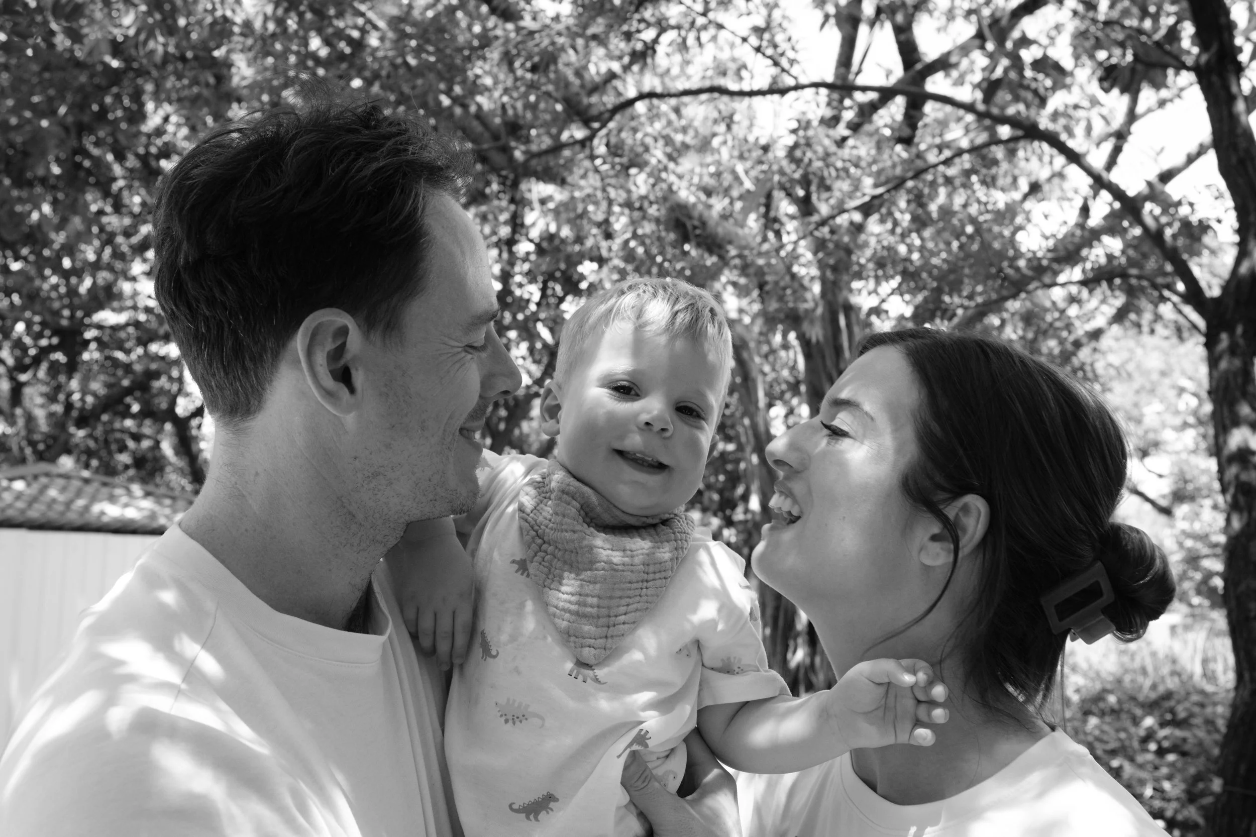 A man and woman are holding their toddler son between them and smiling at him as he looks at the camera and laughs during an outdoor Taipei family photoshoot at Daan Park.