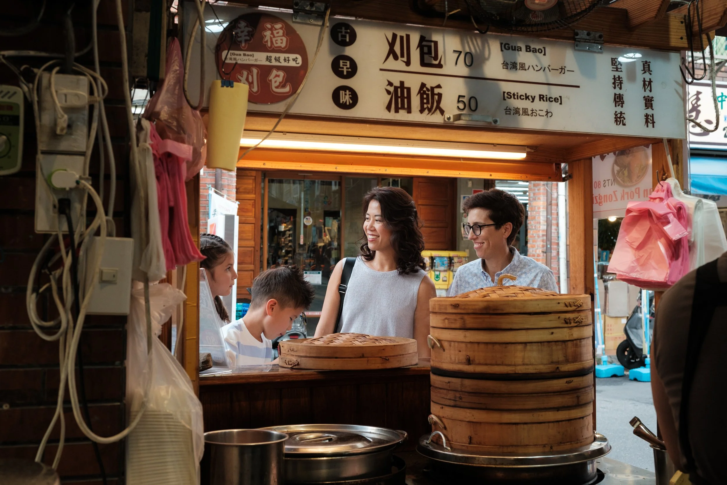 A same-sex family stops at a gua bao stall on Dihua Street, two moms smiling at each other across a counter of bamboo steamers. Taipei family photography.