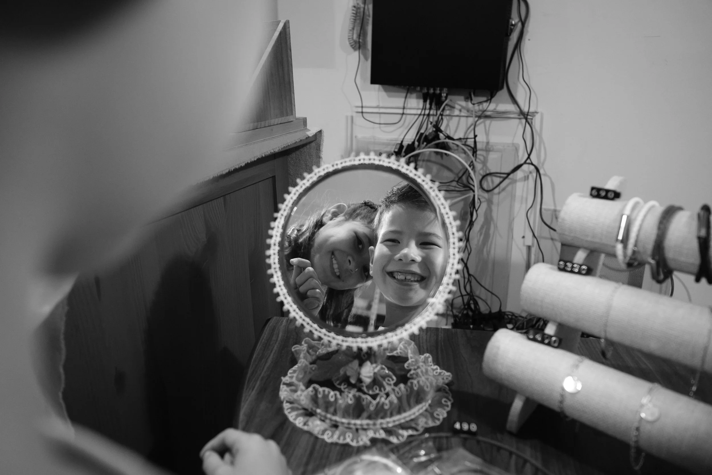 A sister and brother grin at their reflections in an ornate hand mirror inside a Dihua Street shop. Black and white. Taipei family photoshoot.