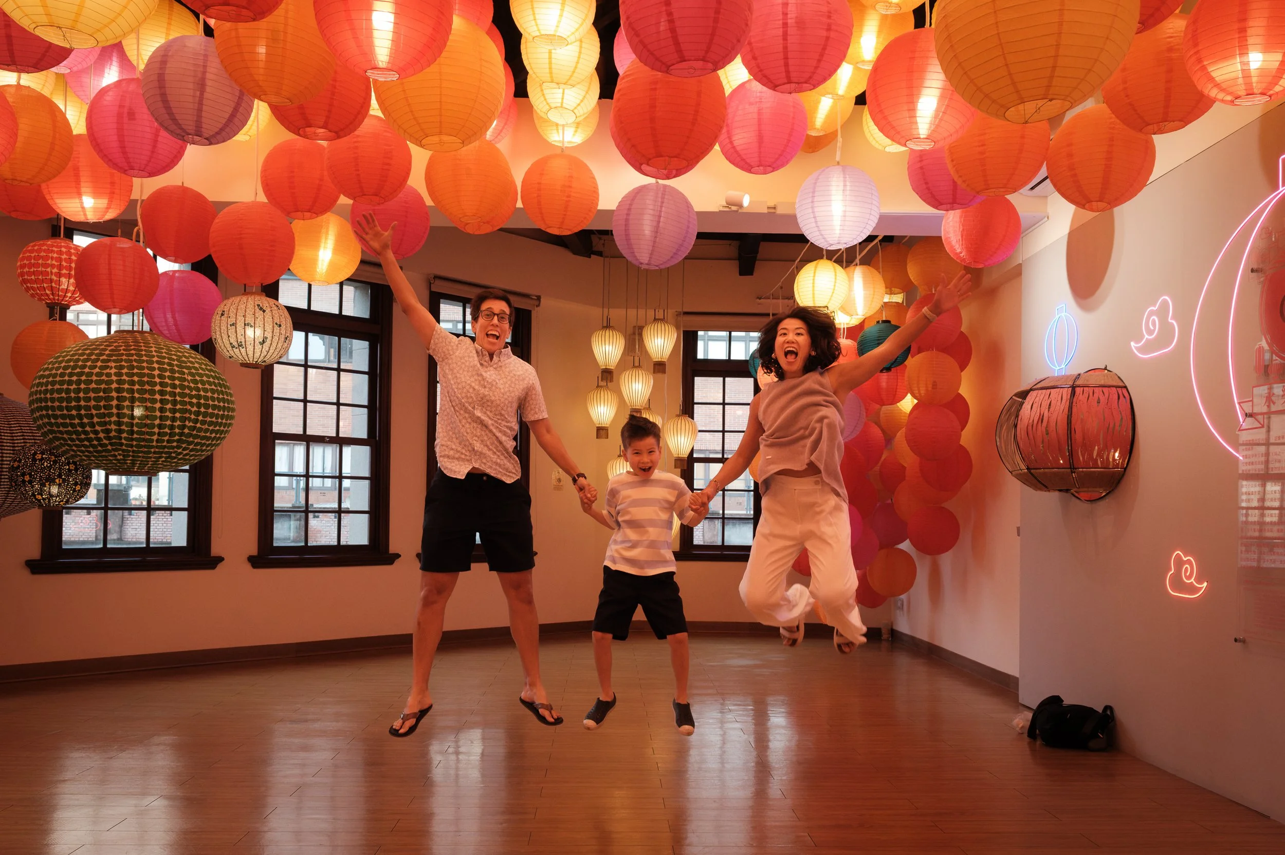 Two moms and their son jump with arms raised inside a room at Dihua Street hung with hundreds of glowing paper lanterns in red, orange, and gold. Taipei family photography.