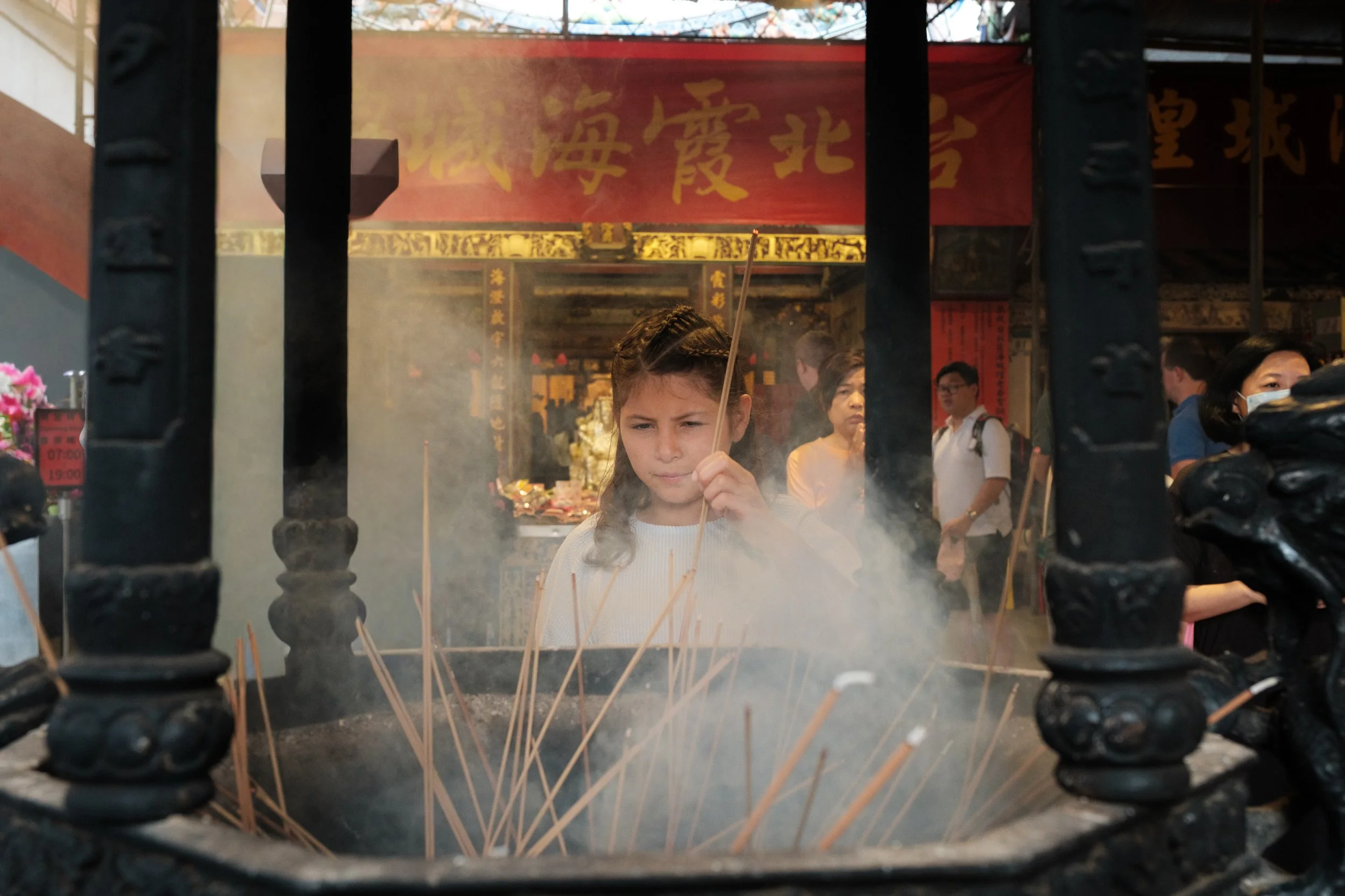 A girl holds a lit incense stick at a Dihua Street temple, smoke billowing around her, focused and still. Documentary family photography, Taipei.