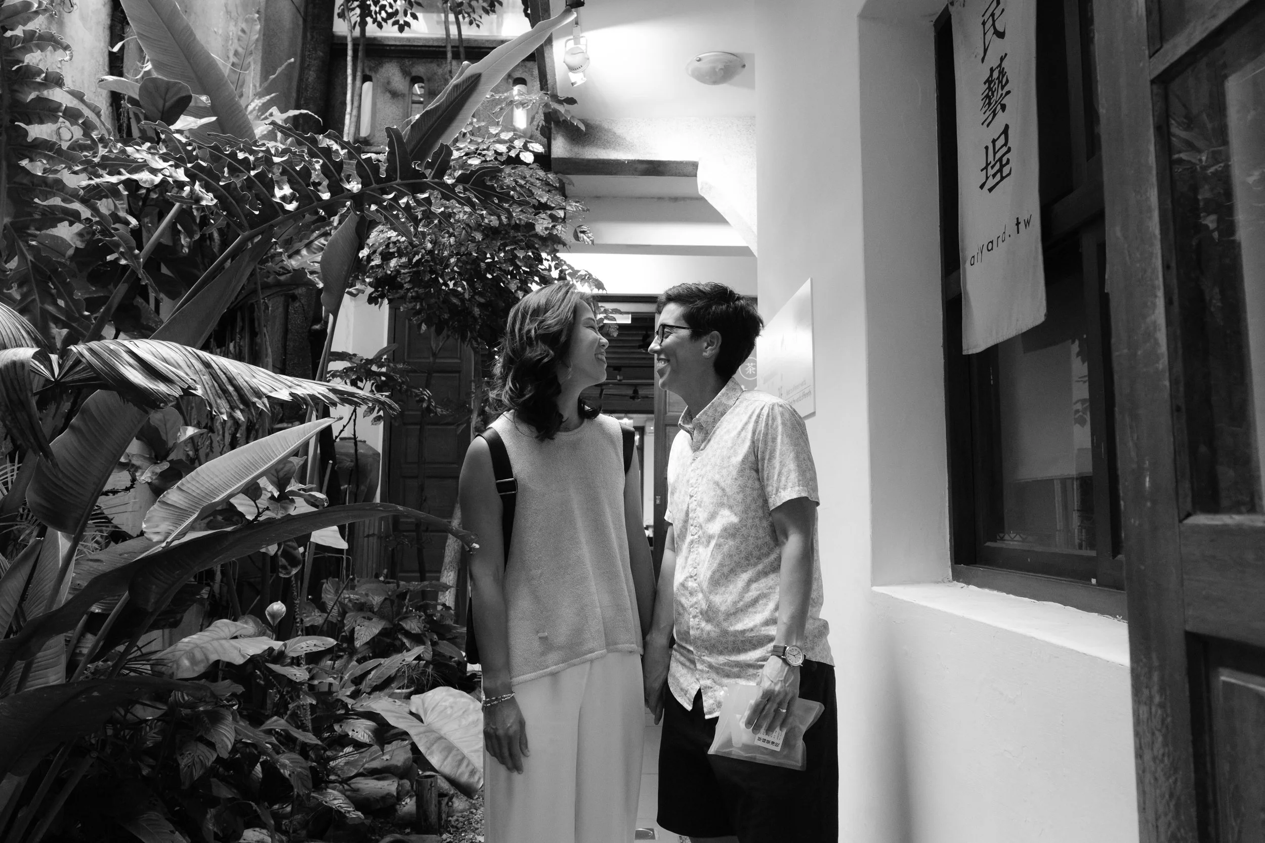 Two moms face each other laughing in a plant-filled Dihua Street courtyard. Black and white. Documentary couples photography, Taipei.