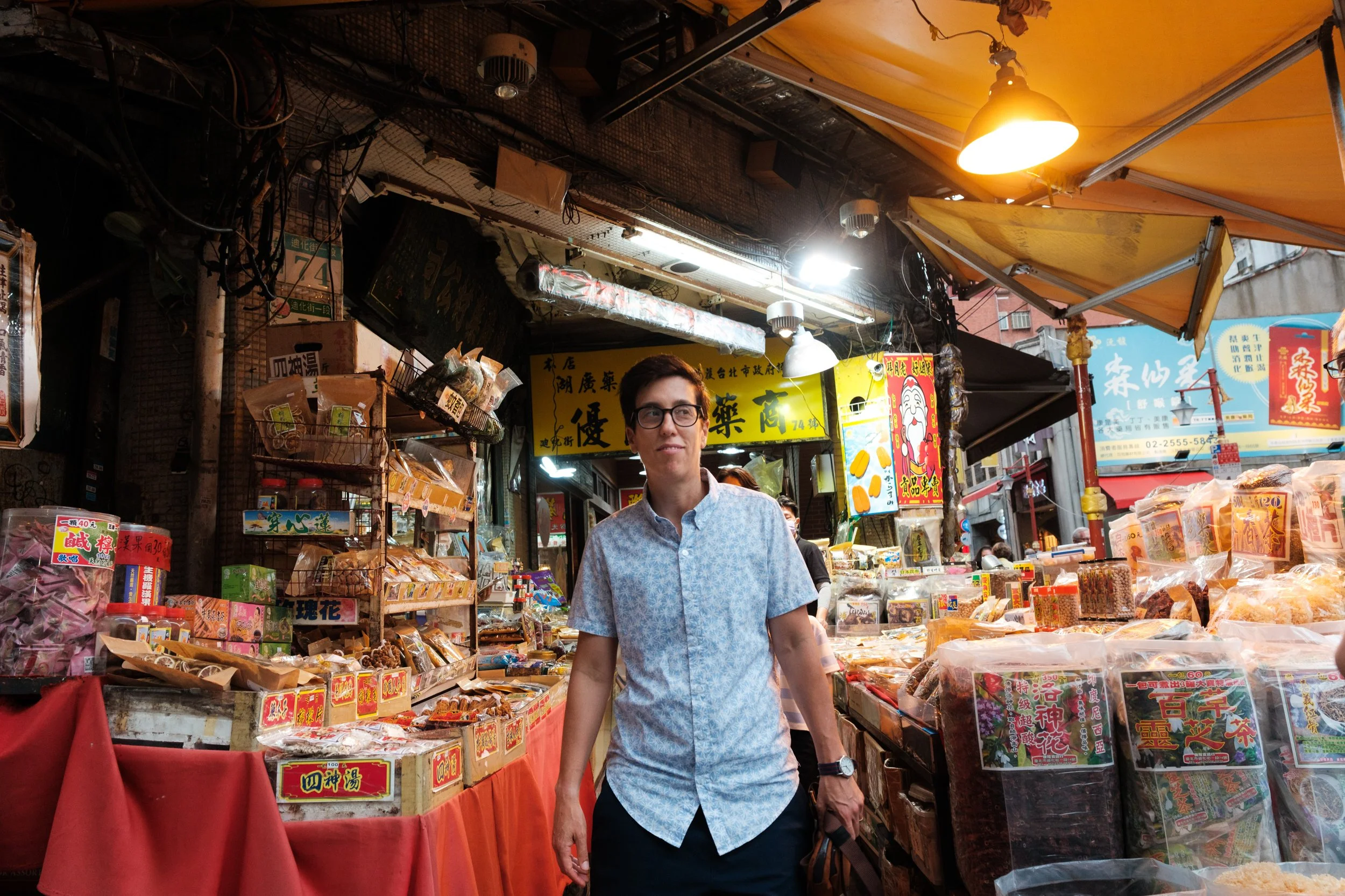 A mom browses a packed Dihua Street market stall of dried goods and herbs under warm yellow light. Taipei outdoor family photoshoot.