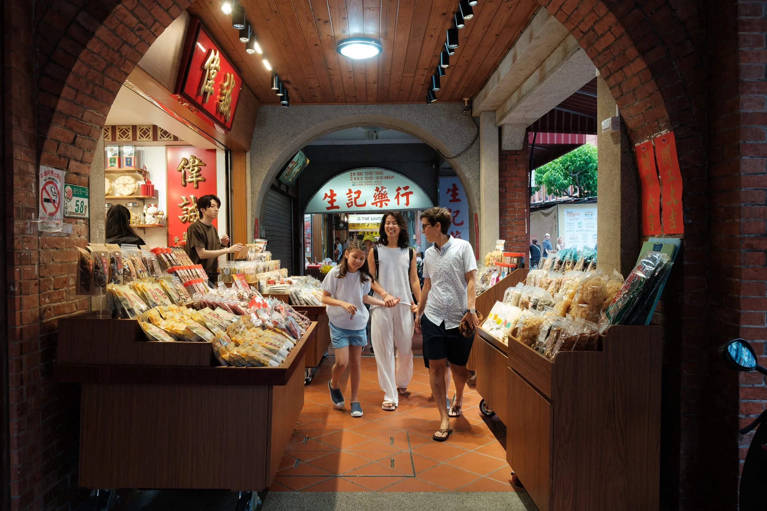 A mom and daughter walk through a covered Dihua Street arcade lined with packaged goods, the other mom just behind. Taipei documentary family photography.