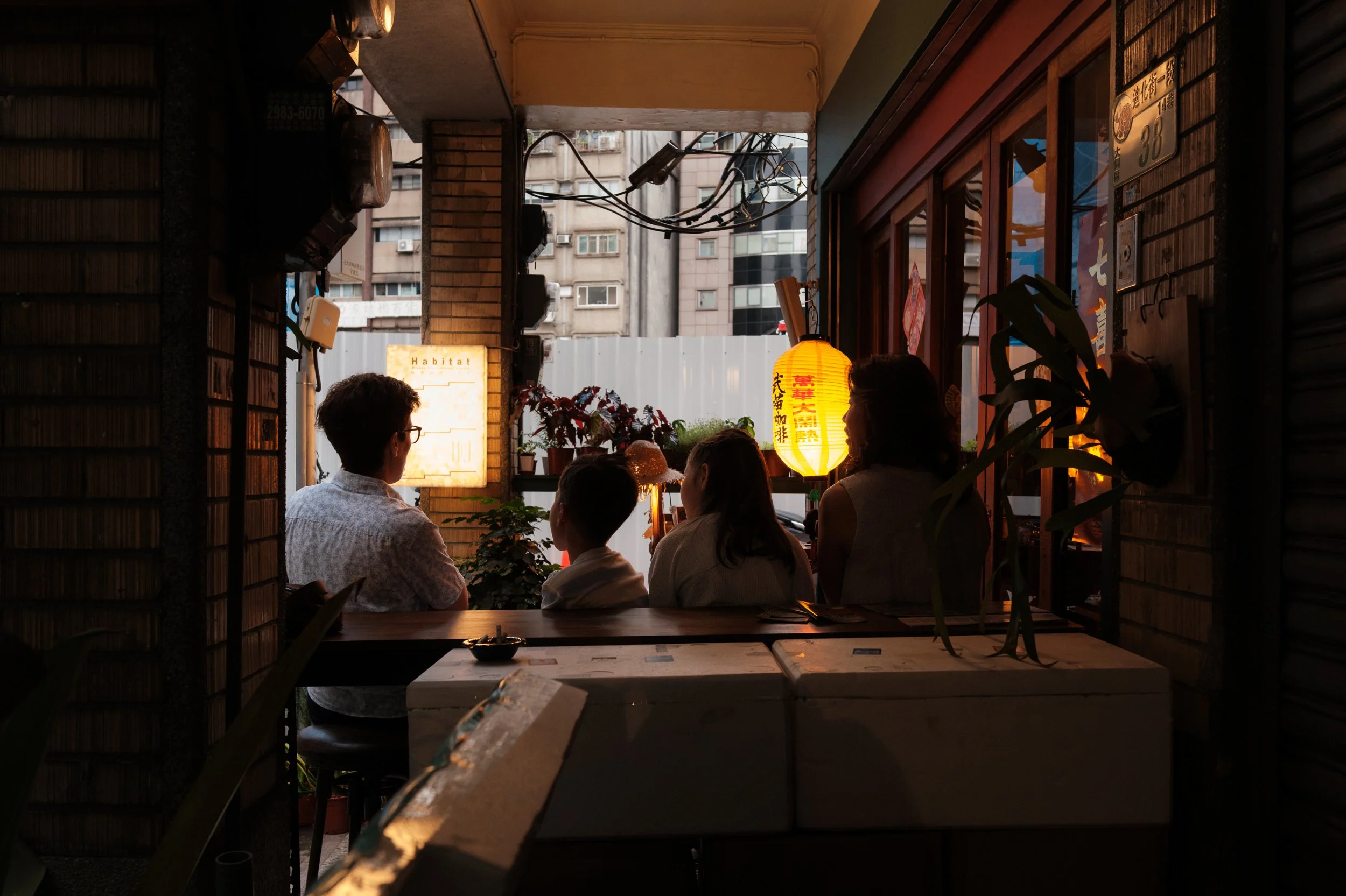 A mom and two kids sit at a café counter on Dihua Street, seen from behind, a glowing paper lantern hanging outside the window. Taipei family photography.