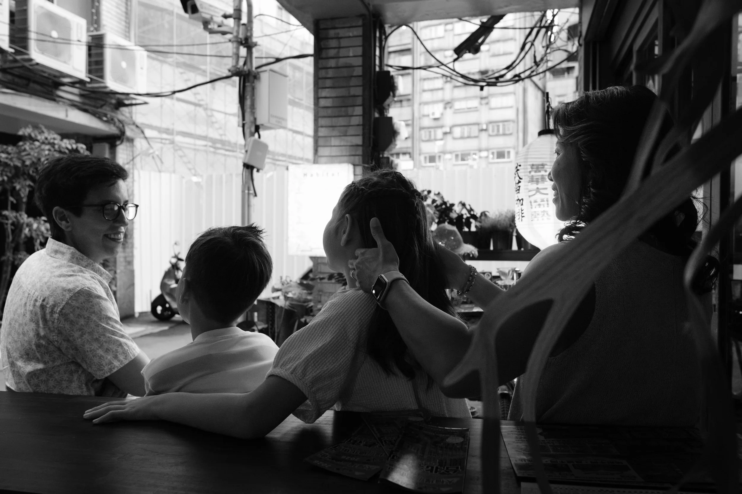 A same-sex family of four sits at a café counter on Dihua Street, one mom stroking her daughter's hair while the other laughs. Black and white. Taipei documentary family photography.