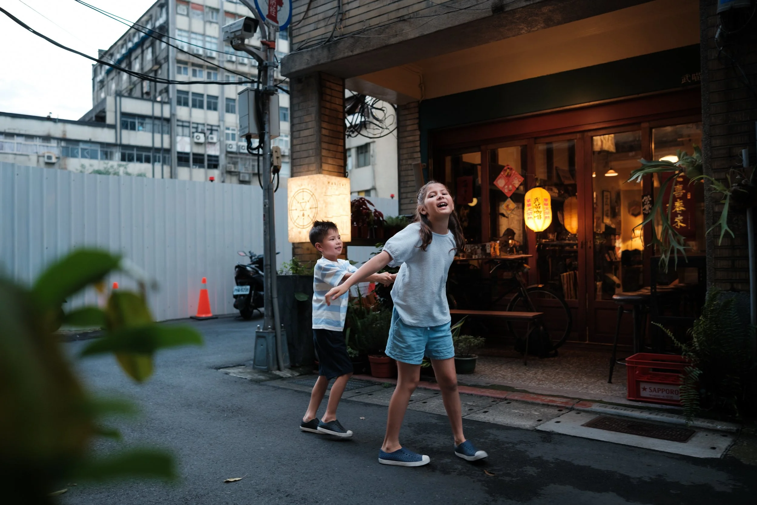 A brother and sister play-fight outside a warmly lit Dihua Street café at dusk, the girl laughing with her head thrown back. Taipei outdoor family photoshoot.