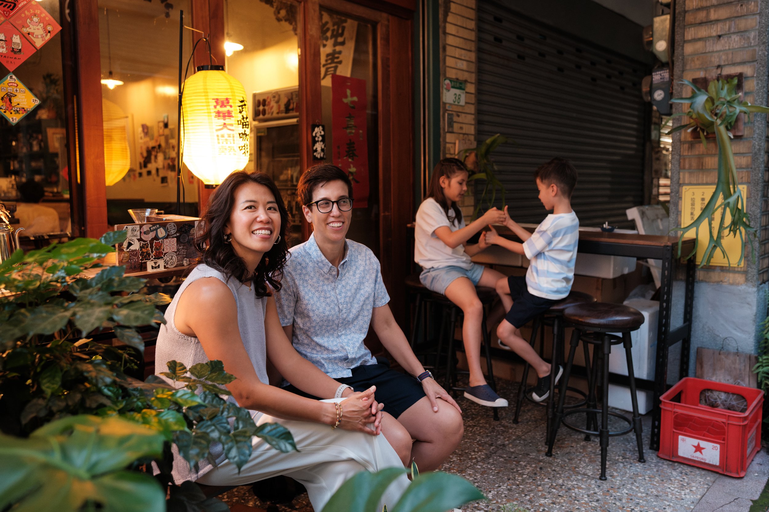 Two moms sit together smiling outside a lantern-lit café on Dihua Street while their kids play on stools behind them. Same-sex family, Taipei family photography.