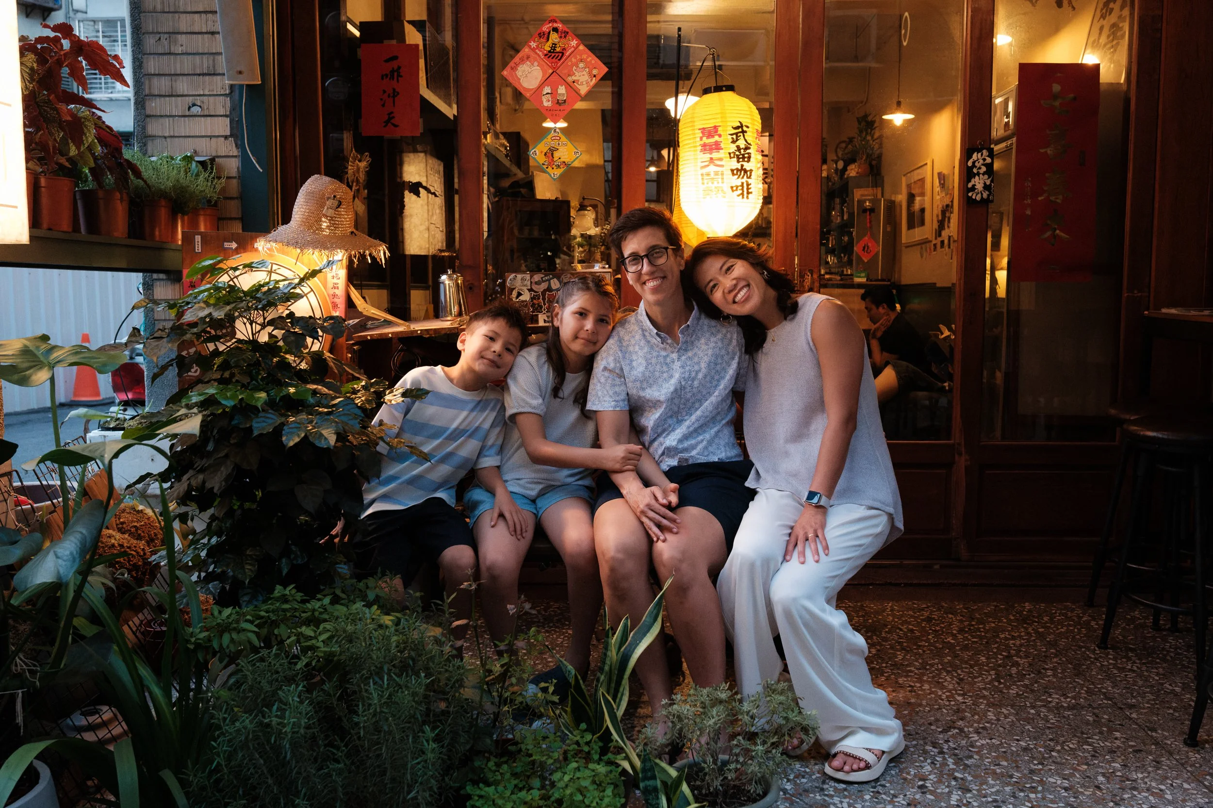 A family of four — two moms and two kids — huddle together laughing outside a glowing lantern-lit café on Dihua Street, Taipei. Outdoor family photoshoot.