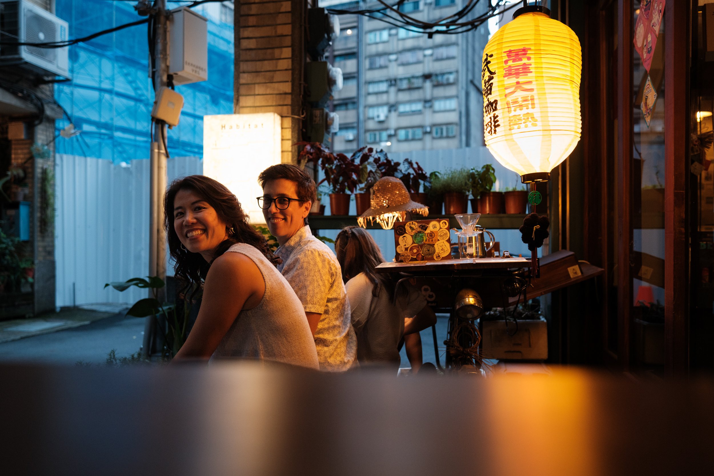 Two moms look back over their shoulders and laugh together outside a Taipei café at dusk, a paper lantern glowing above them. Documentary family photography, Taipei.
