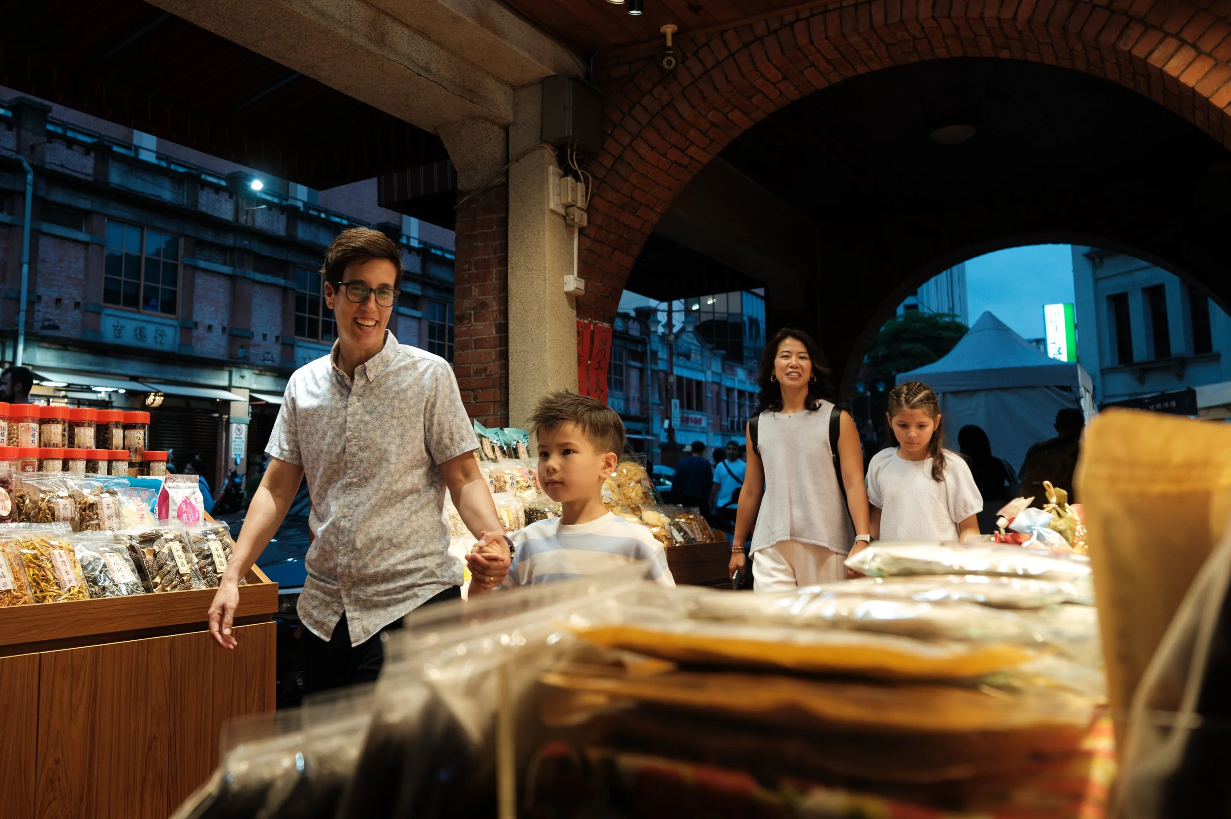 A mom leads her young son by the hand through a Dihua Street market arcade at dusk, the other mom and daughter following behind under brick arches. Taipei family photoshoot.