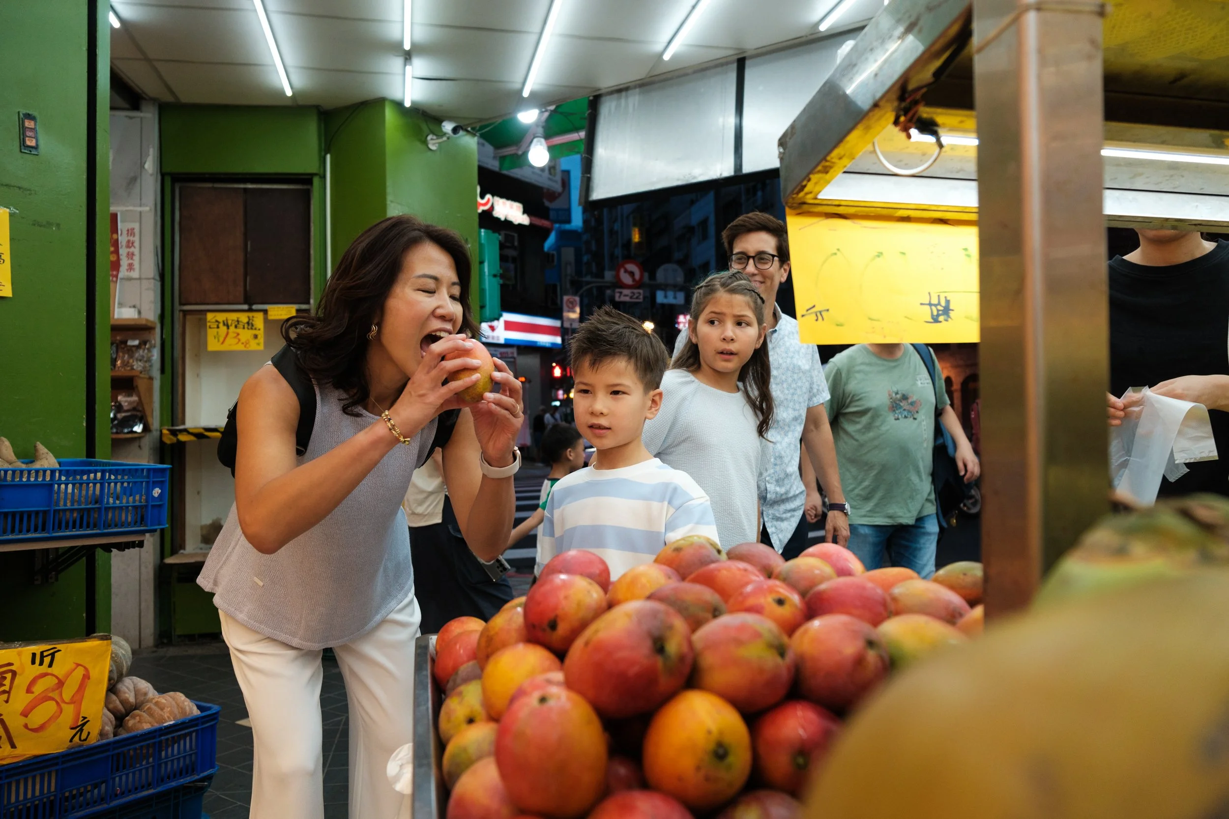 A mom pretends to bite into a mango at a Ningxia Night Market fruit stall while her son watches, the rest of the family visible in the busy market behind them. Taipei family photography.