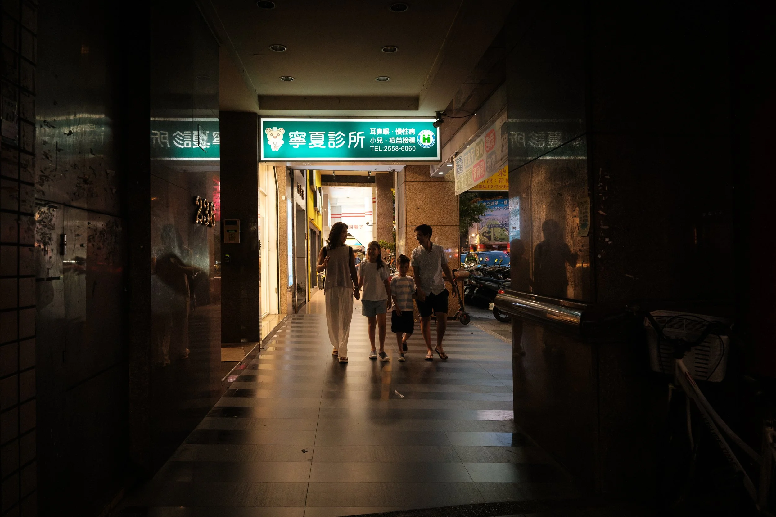 A family of four walks hand-in-hand in silhouette through a dark Taipei arcade passage toward a glowing green clinic sign, the city street visible beyond. Documentary family photoshoot, Taipei.